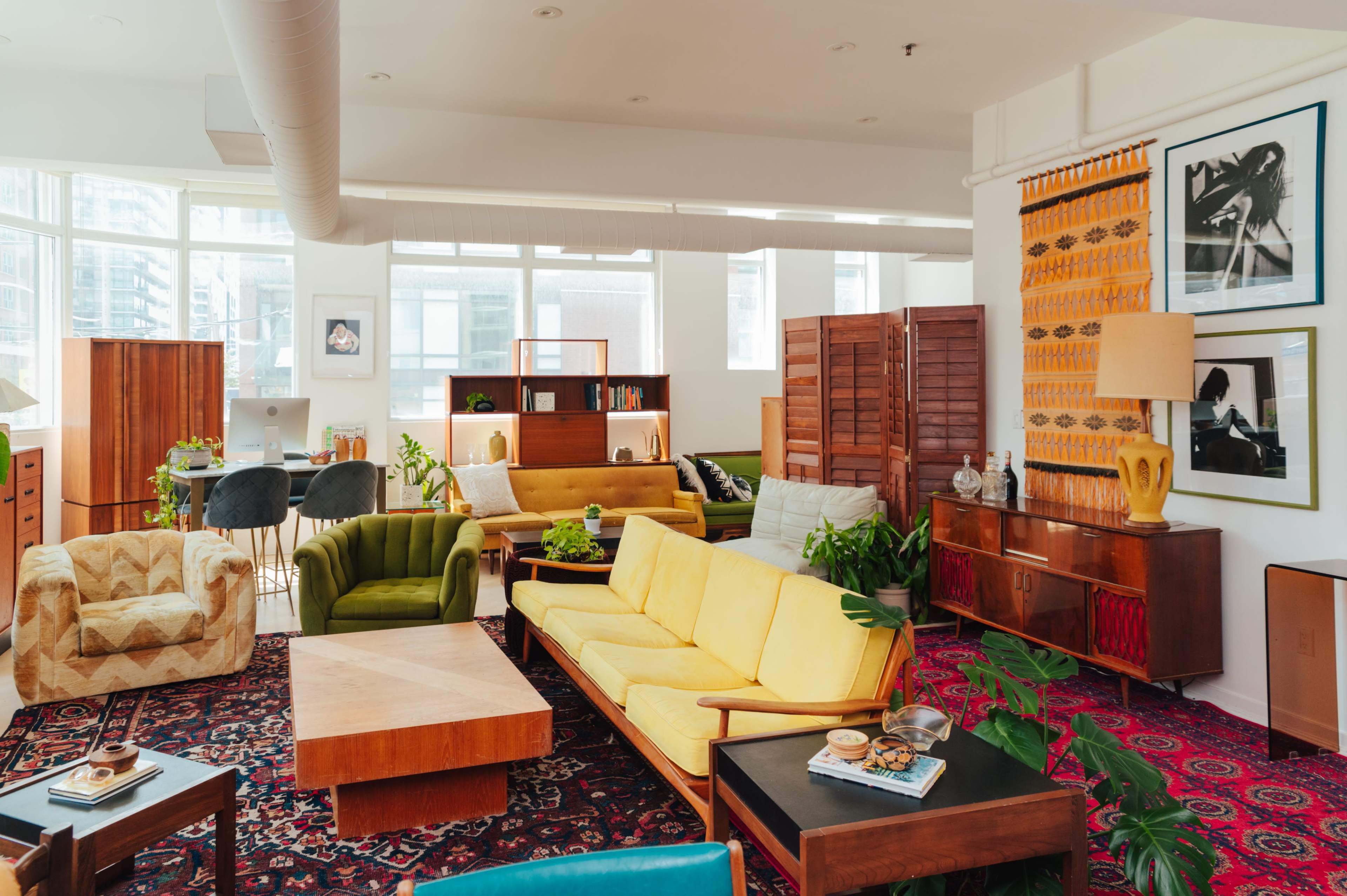 A spacious, well-lit living room featuring various vintage furniture pieces, plants, and decorative items arranged around a colorful area rug.