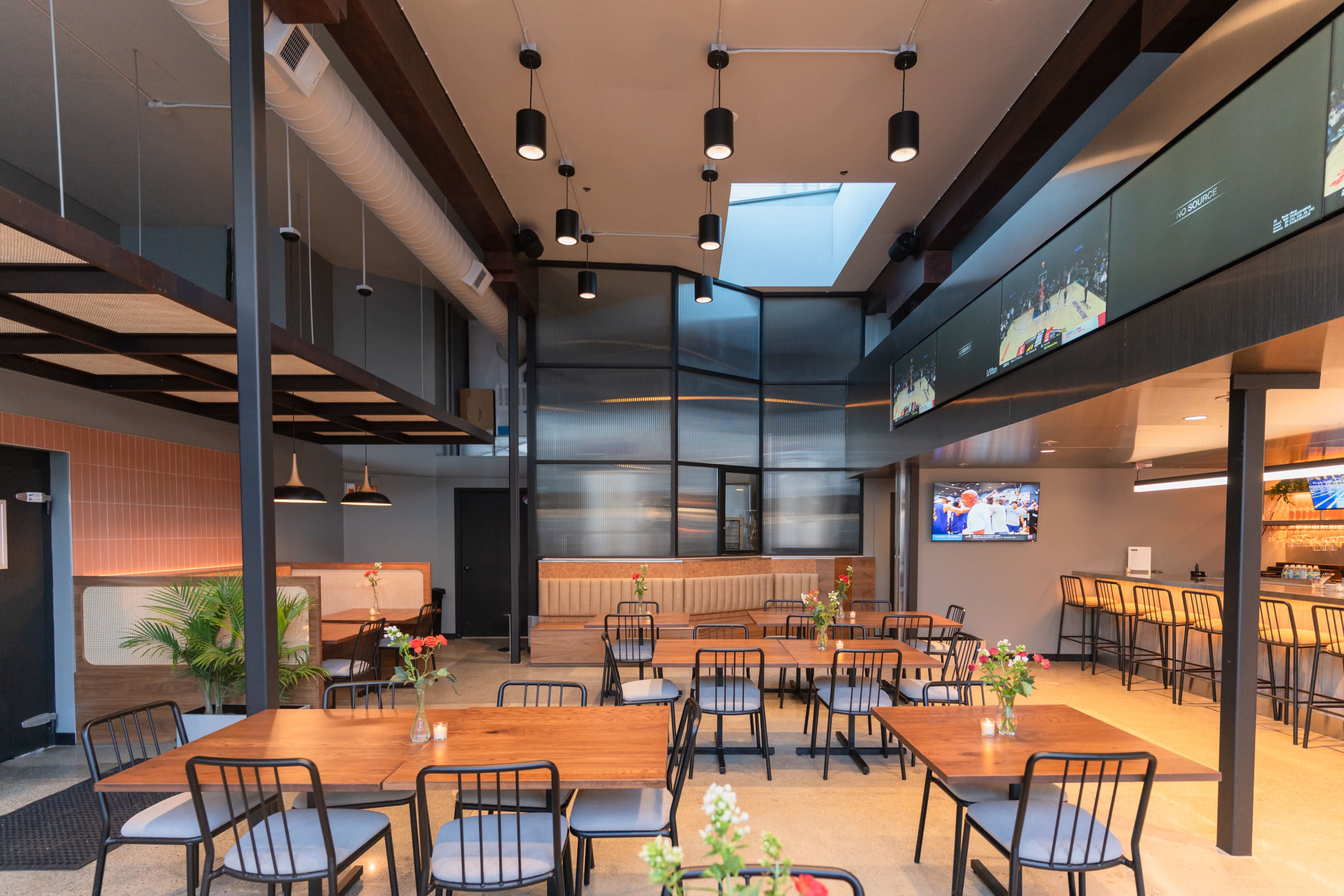 The image shows a modern dining area with wooden tables and black chairs, featuring large windows and screens displaying sports events.