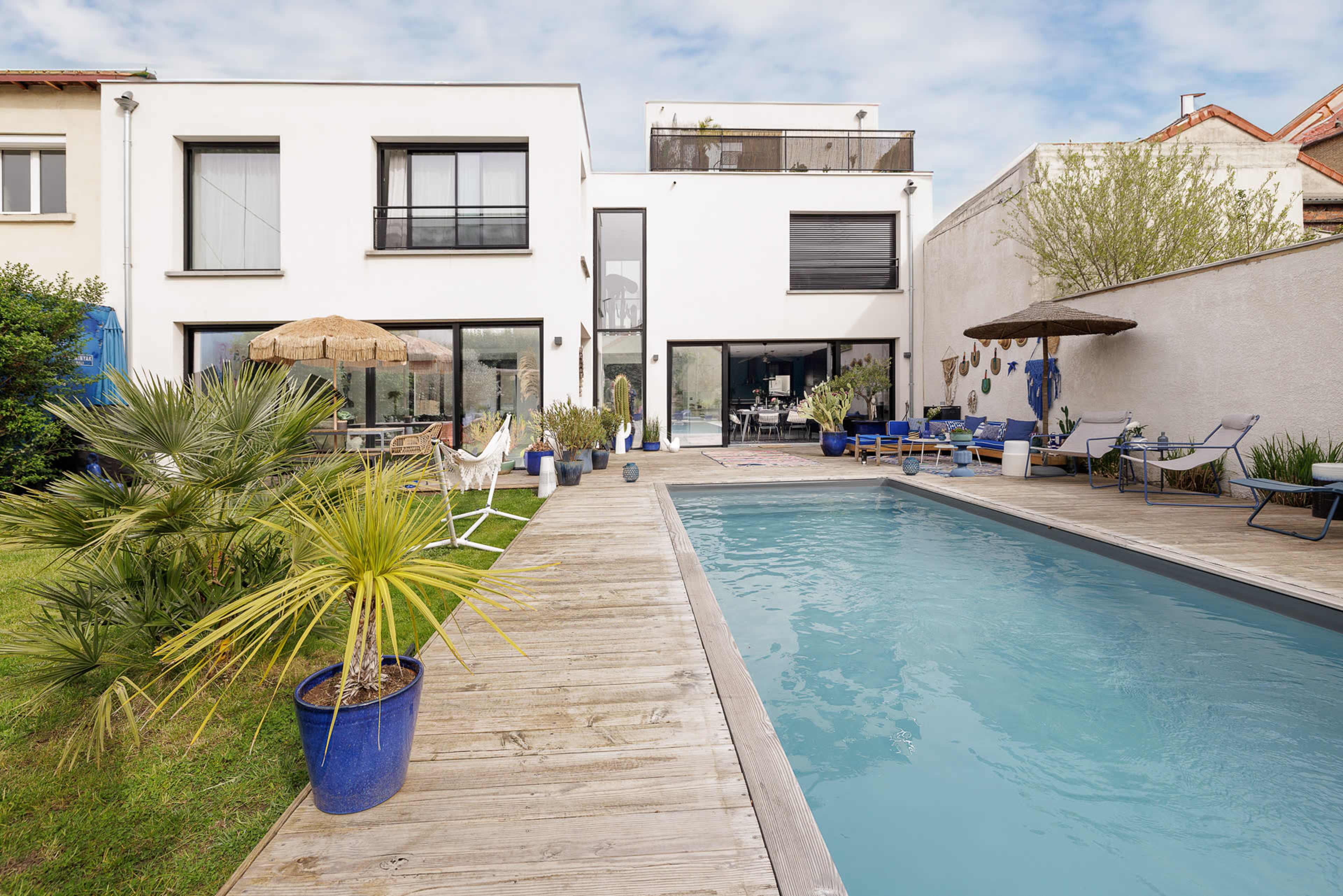 A modern two-story house with a swimming pool lined by wooden decking and surrounded by lush greenery and patio furniture.