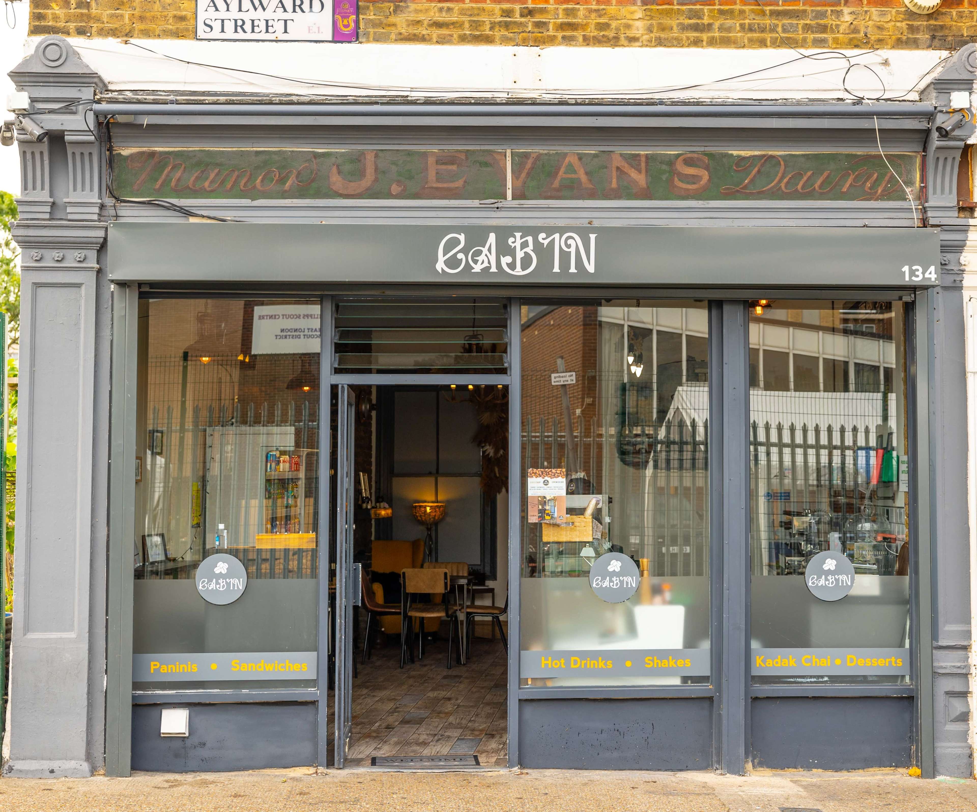The storefront of a café named "CABIN," featuring large glass windows and a sign that includes the words "Paints & Sandwiches" and "Hot Drinks + Shakes + Kodak Eats & Desserts."