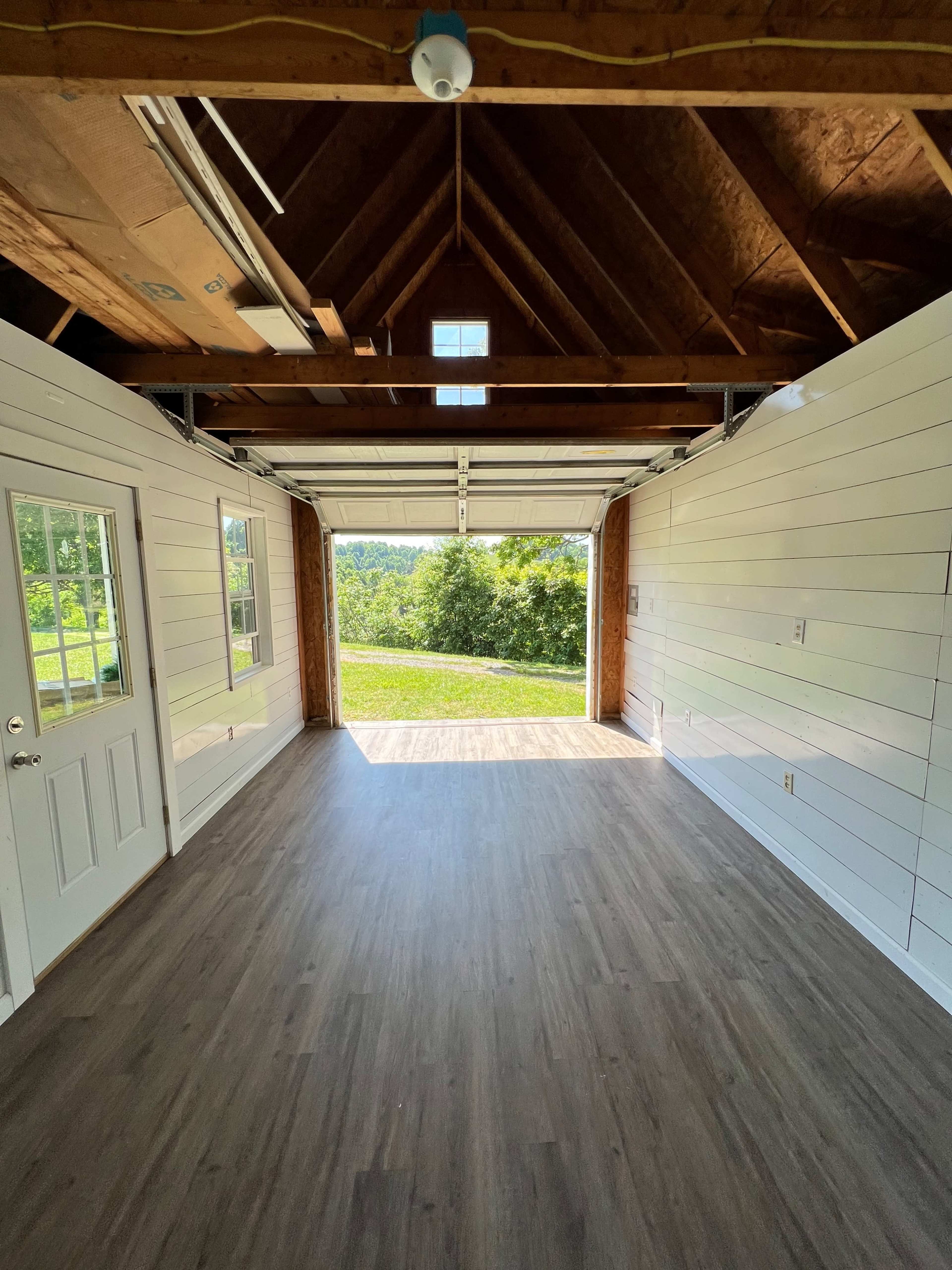 The image shows the interior of a spacious garage with a gabled ceiling, featuring wooden paneling and large doors opened to a view of greenery outside.