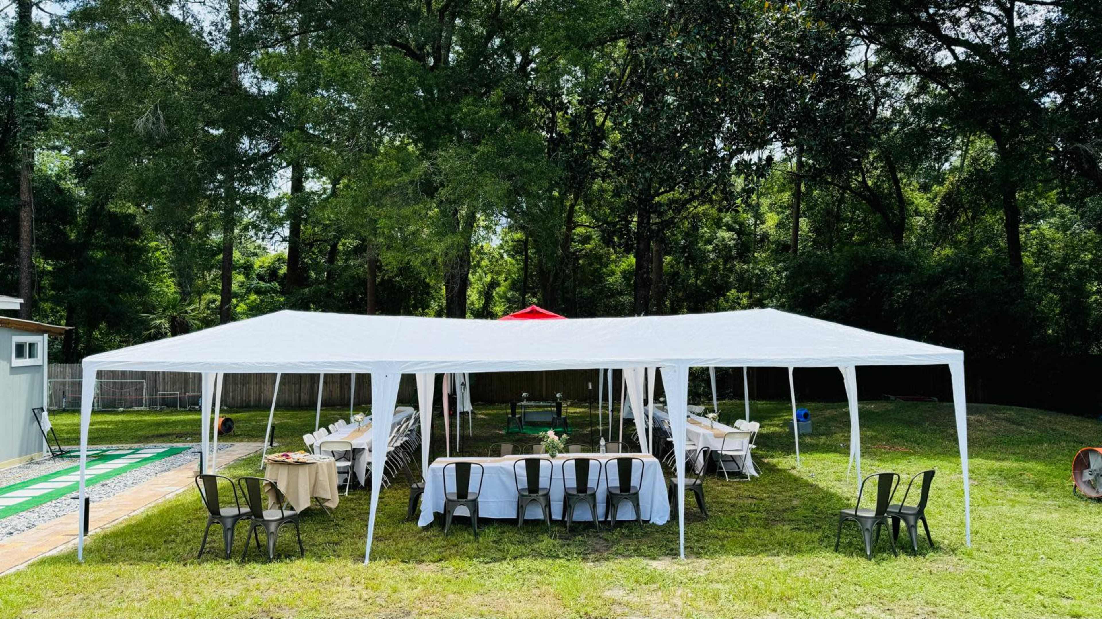 A white tent is set up in a grassy area, with tables arranged underneath for a gathering, surrounded by trees.