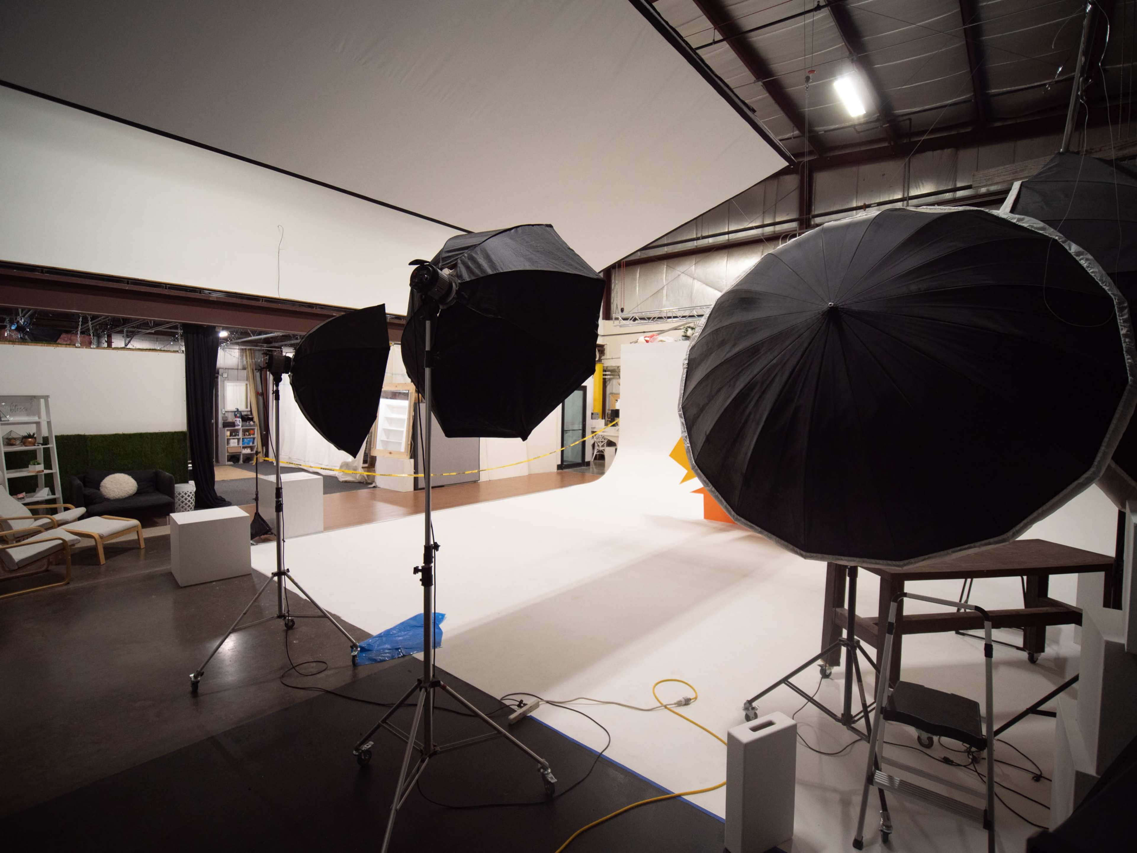 A photography studio with three large softbox lights positioned on stands, illuminating a white backdrop and various props in the background.