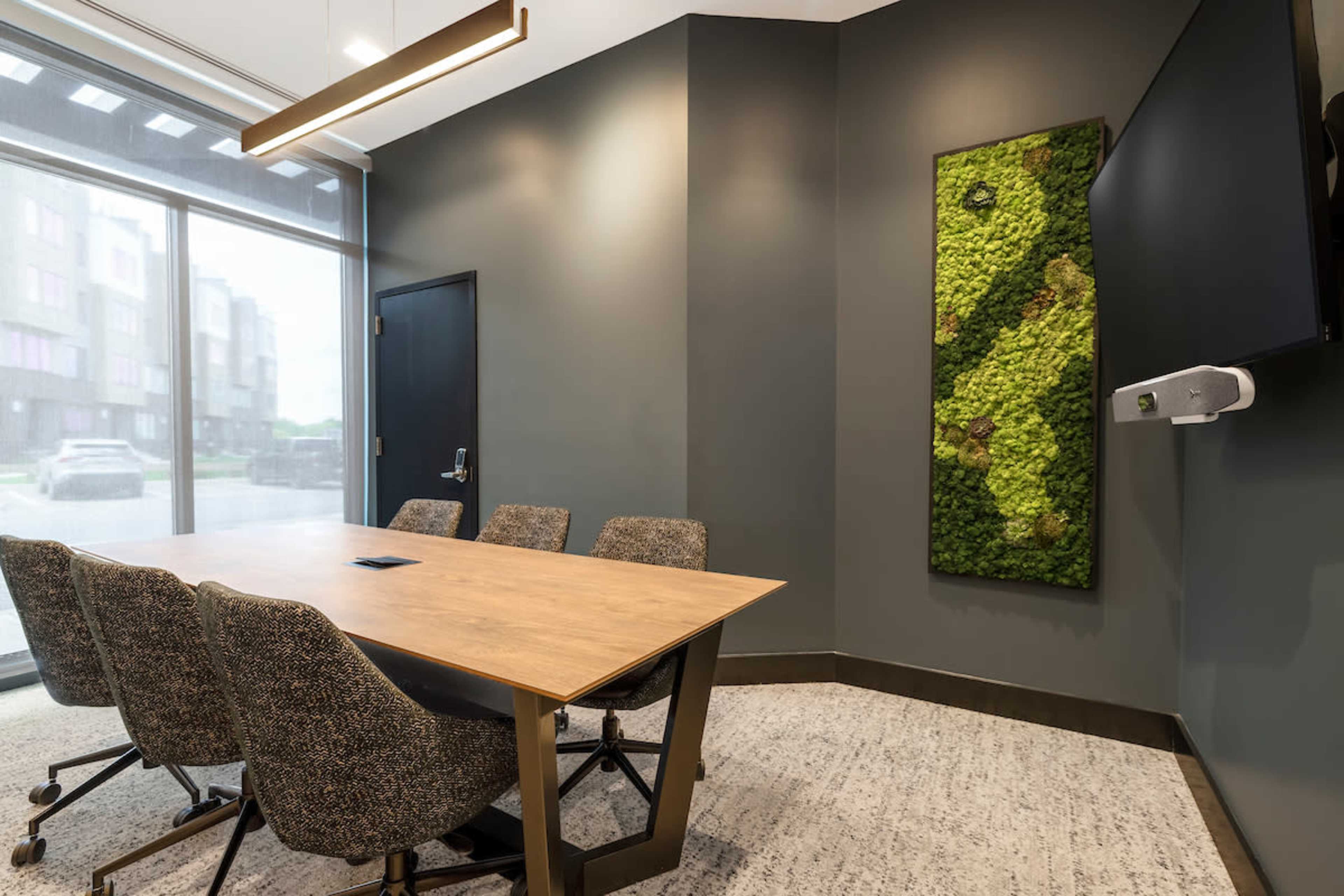 A conference room features a wooden table surrounded by four upholstered chairs and a green moss wall art on the gray wall.