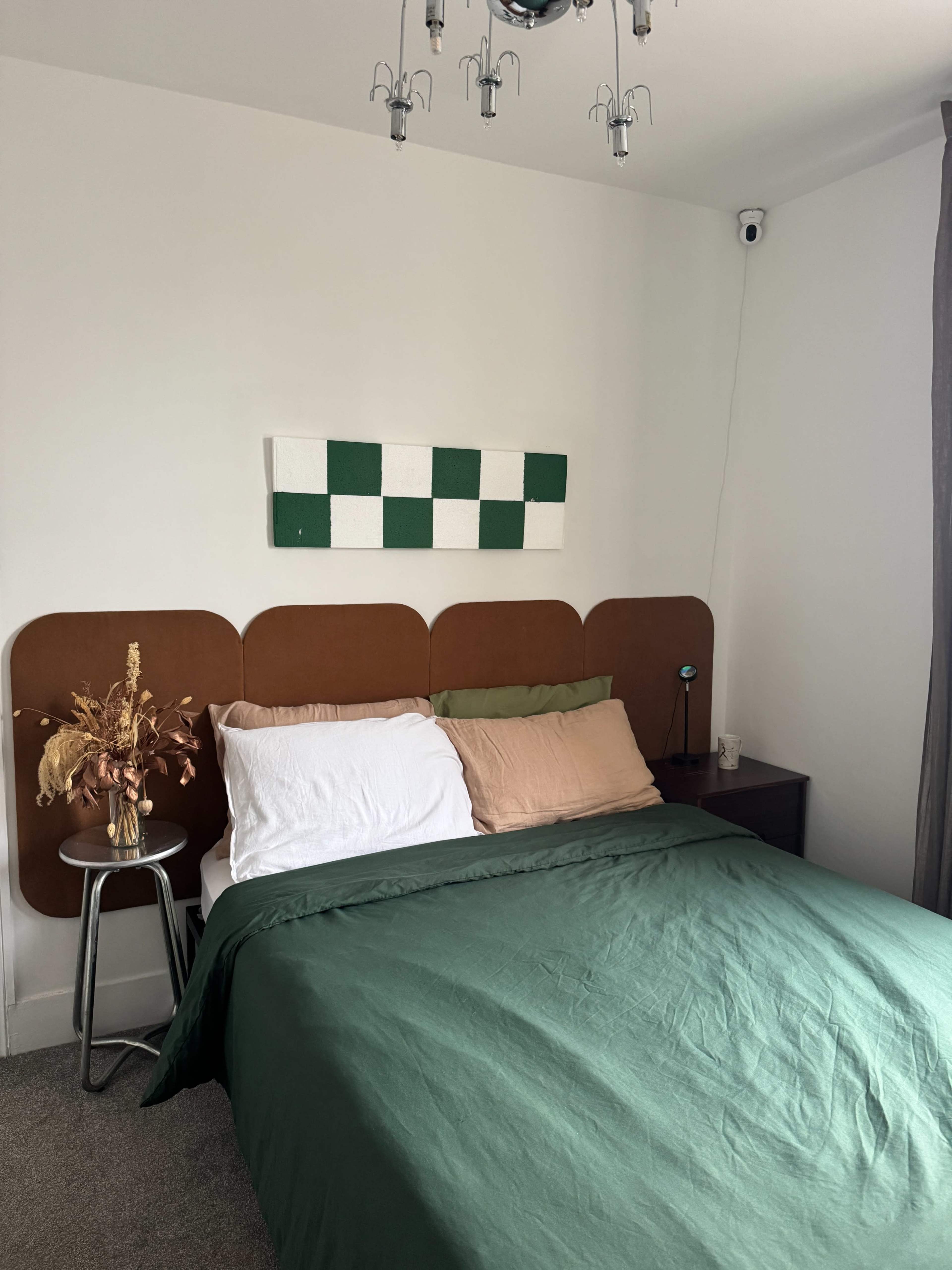 The image shows a bedroom with a double bed covered in a green duvet, two contrasting pillows, and a decorative wall hanging above the headboard.