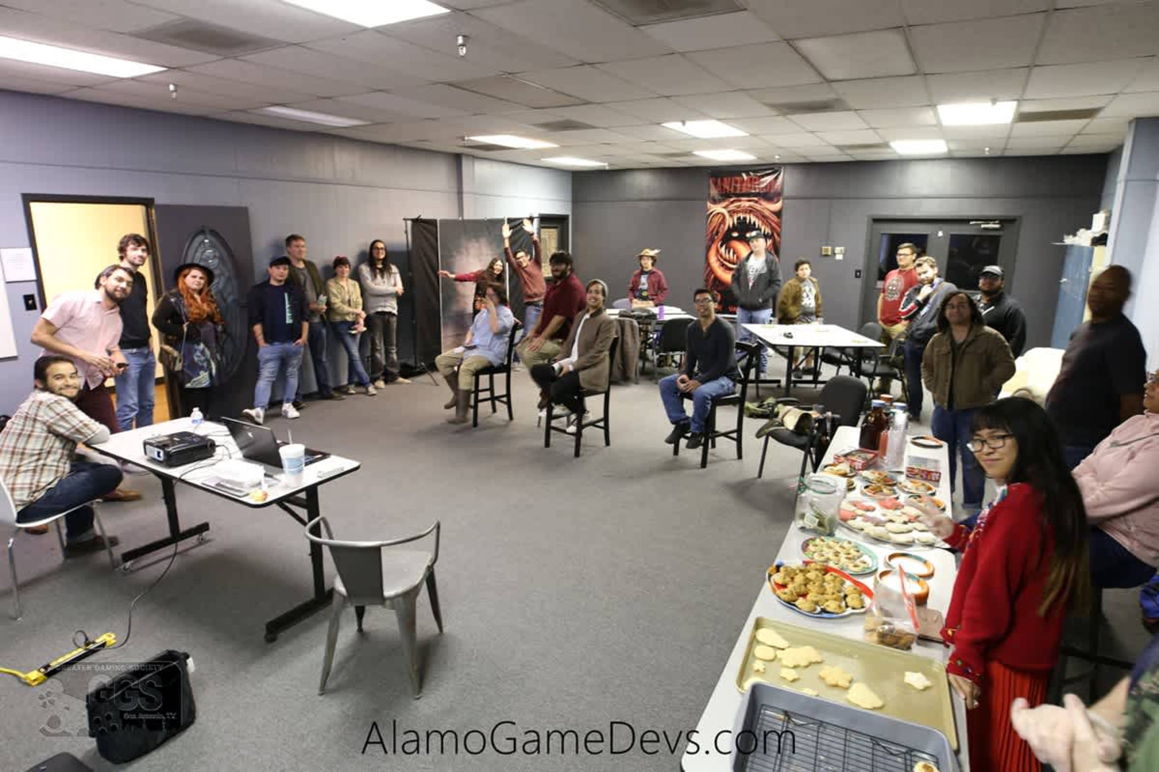 A group of people is gathered in a meeting room with tables set up displaying food, while some attendees are seated and others are standing.