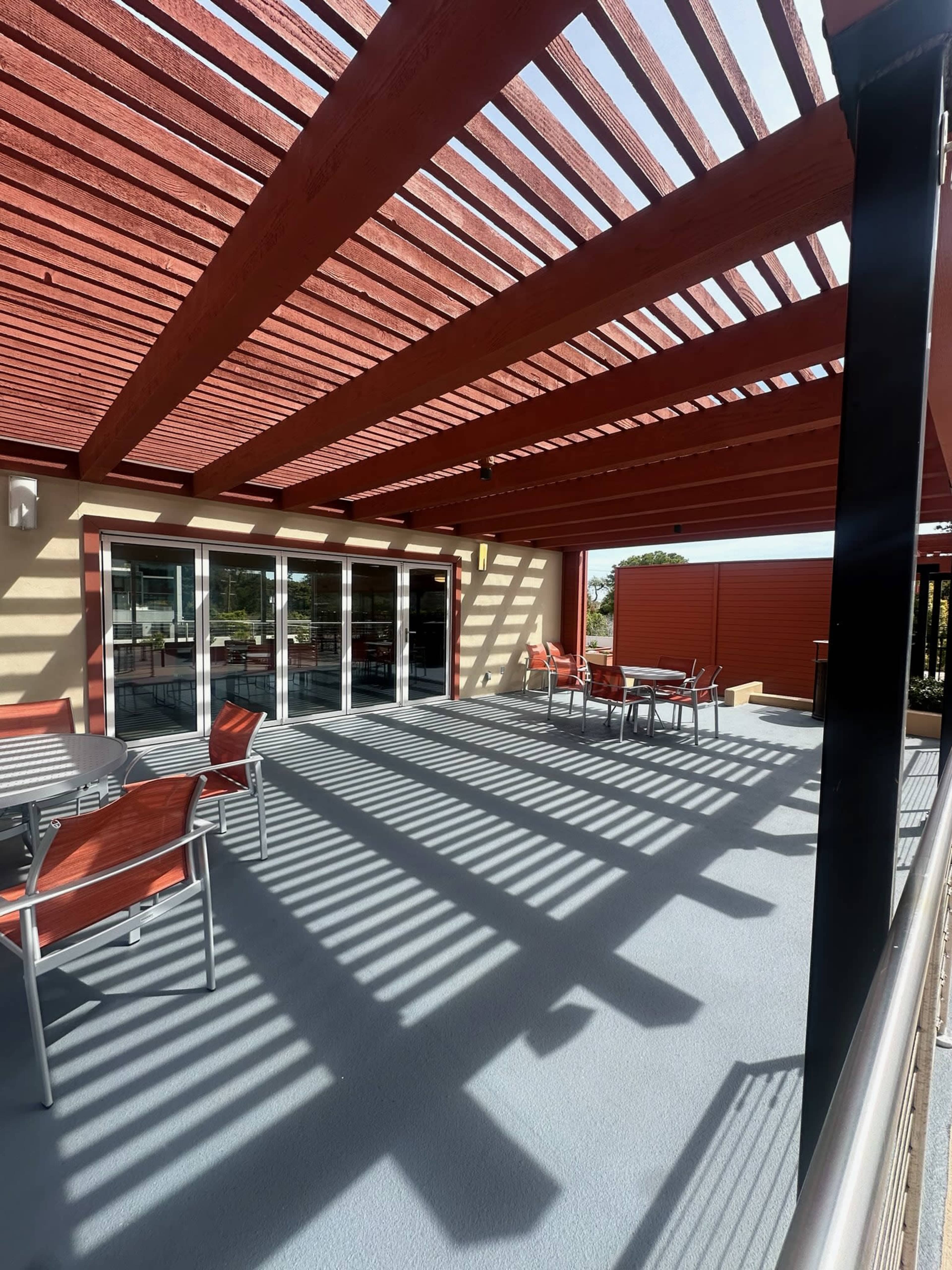 The image shows a spacious outdoor patio area with a wooden pergola casting shadows over metal tables and chairs under blue skies.