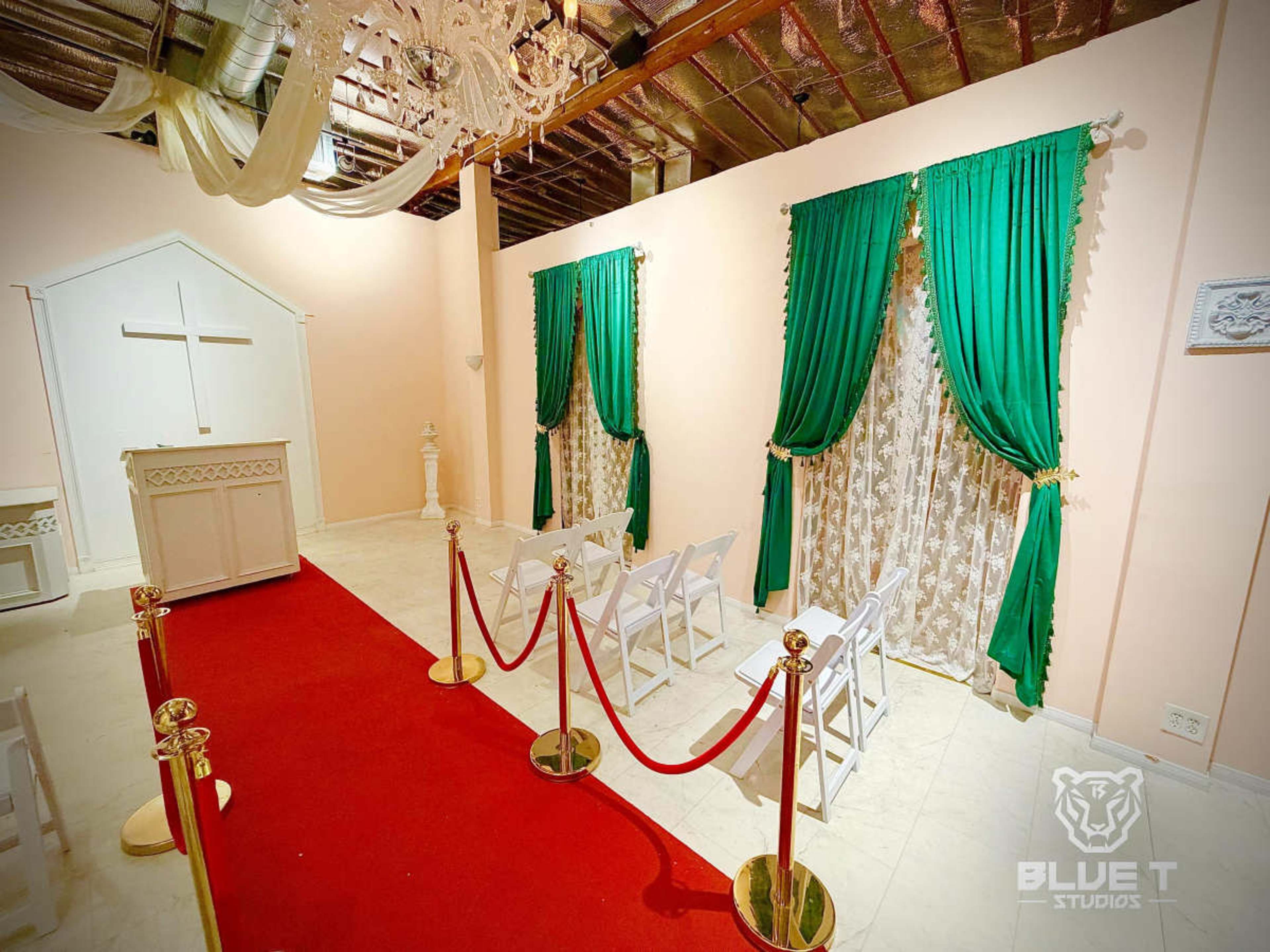 The image shows a room set up for a ceremony, featuring a red carpet, white chairs, and green drapes framing windows, with a small altar at one end.