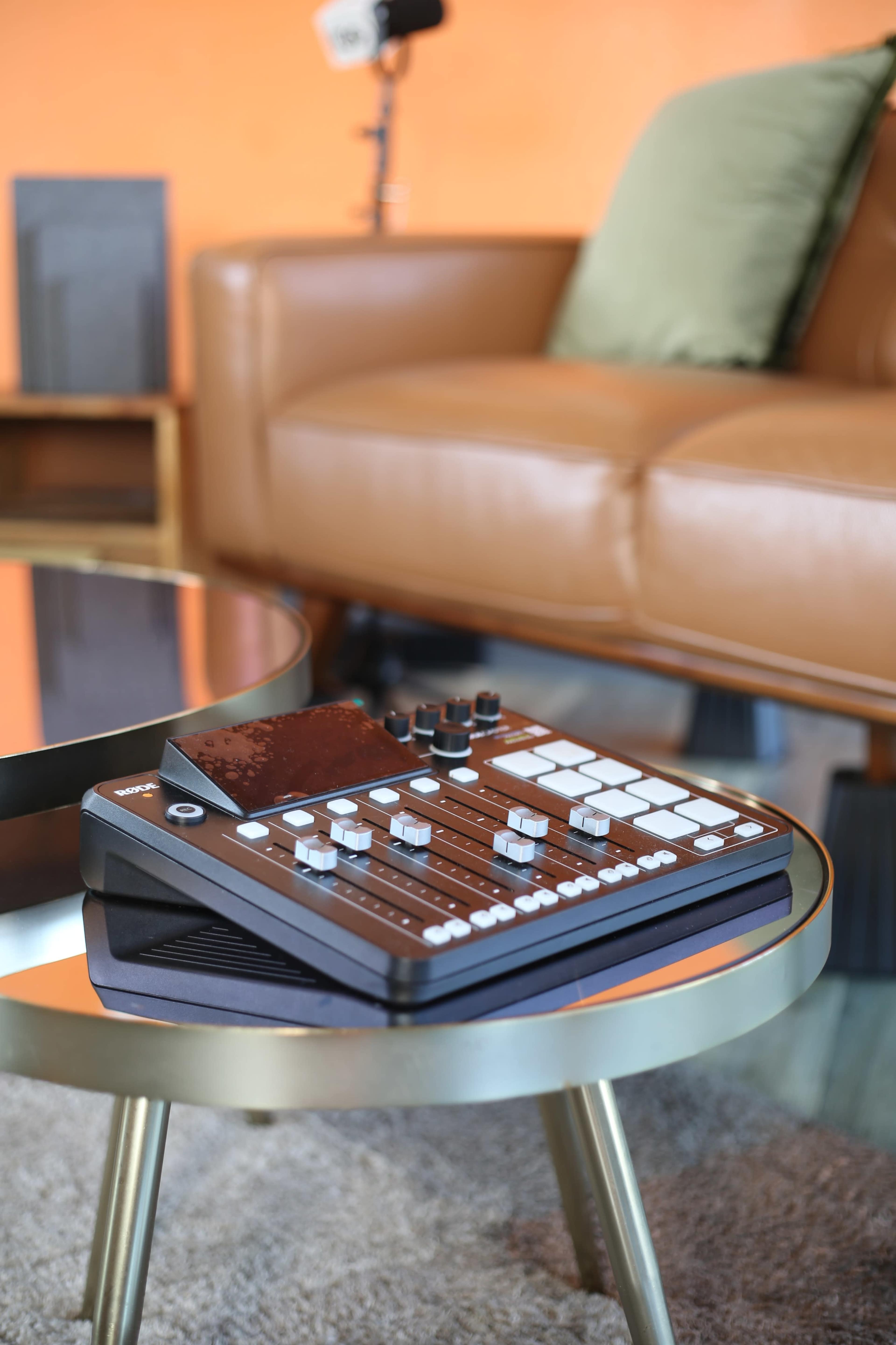 A music production controller lies on a round coffee table in a cozy living room setting with a leather sofa and an orange wall.