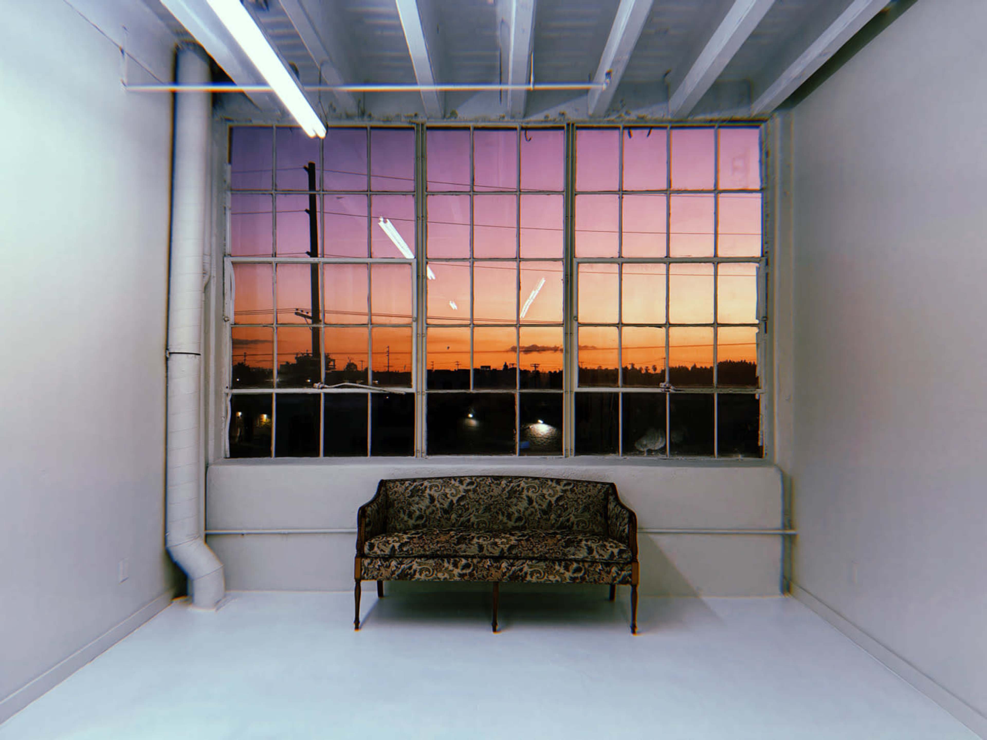 A vintage sofa is positioned in front of large windows that display a vibrant sunset sky.