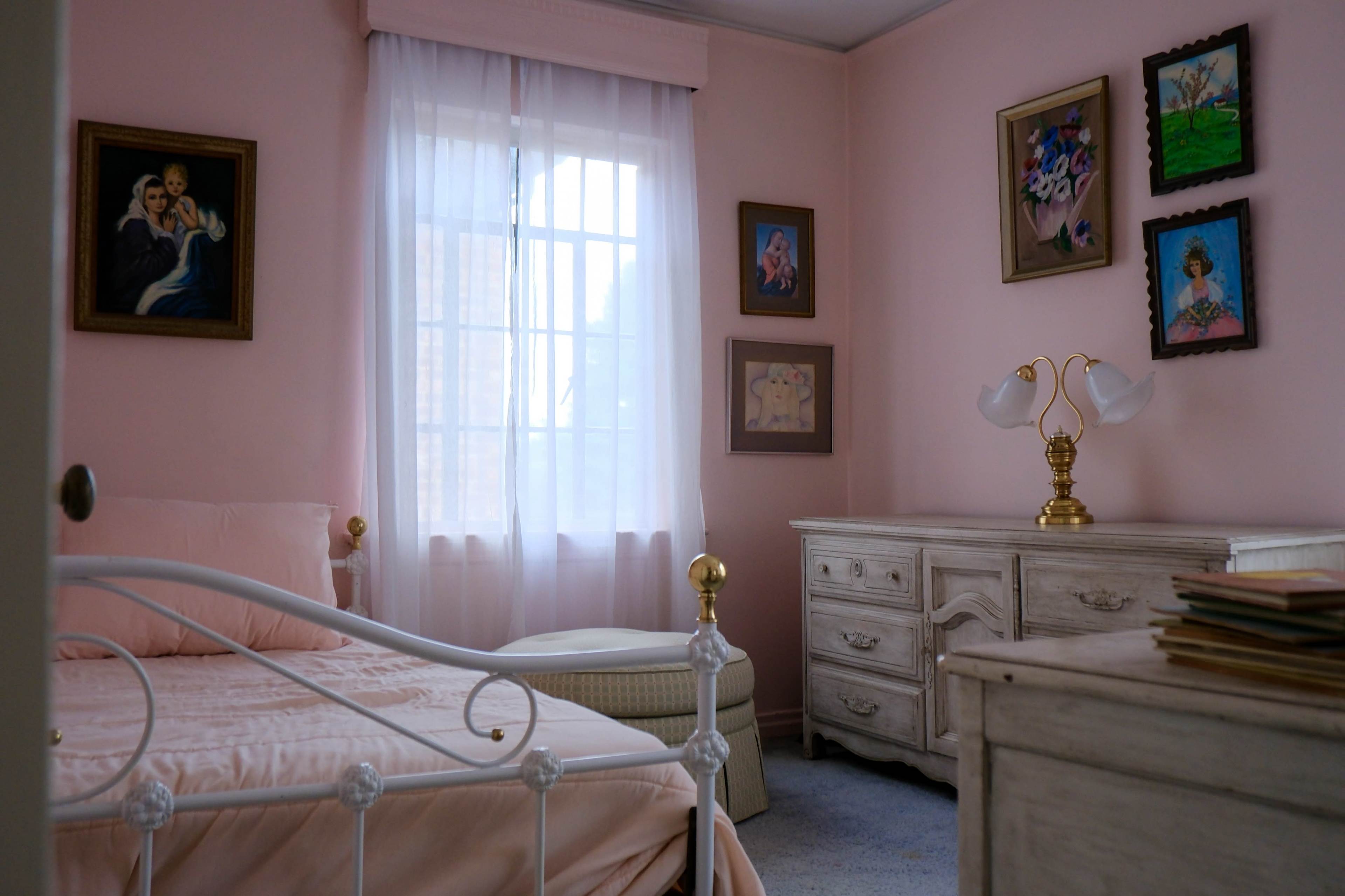 The image shows a softly lit bedroom with pink walls, a white bed, a dresser, and several framed paintings on the walls.