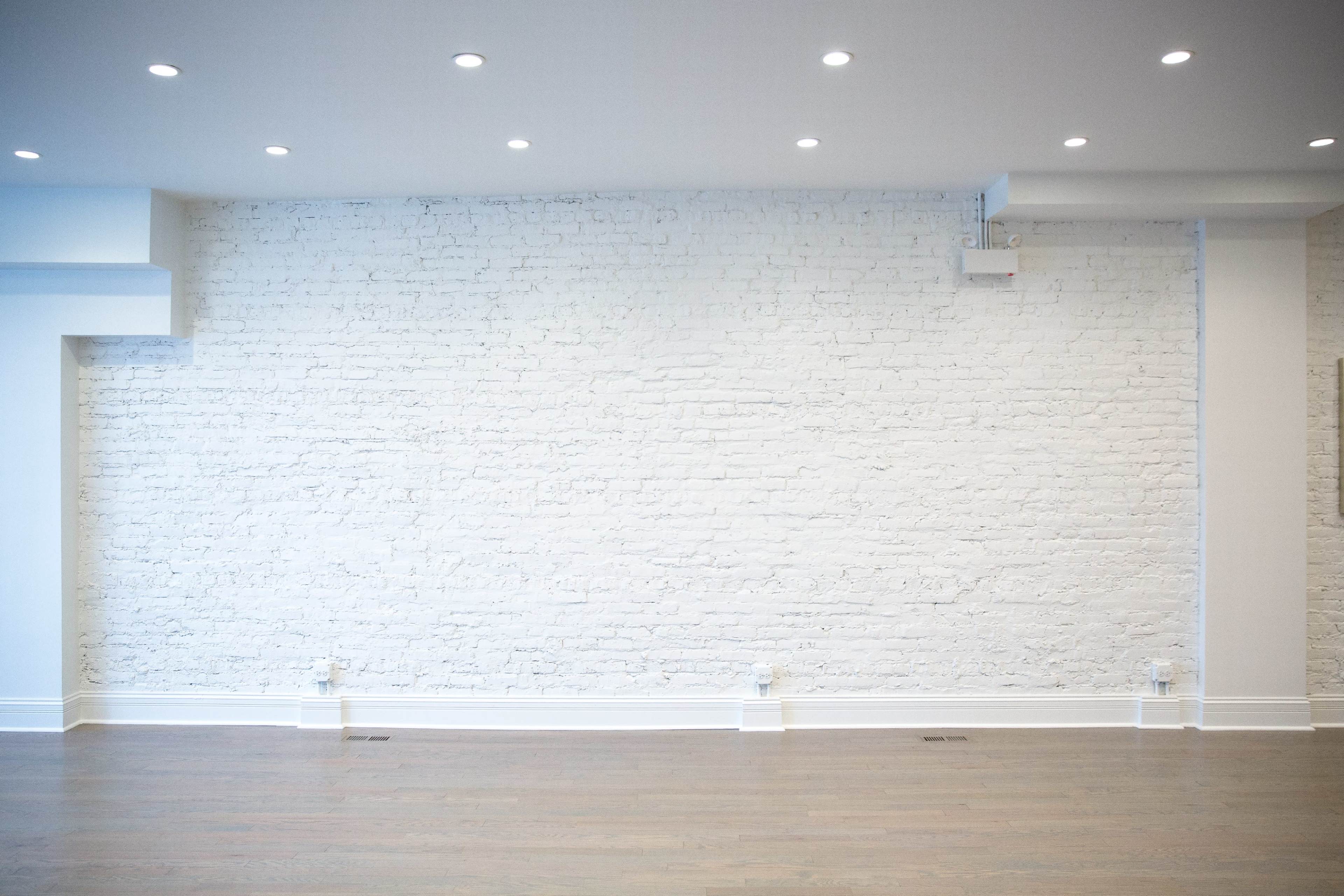 The image shows a spacious room with a white brick wall and wooden flooring, illuminated by recessed lighting.