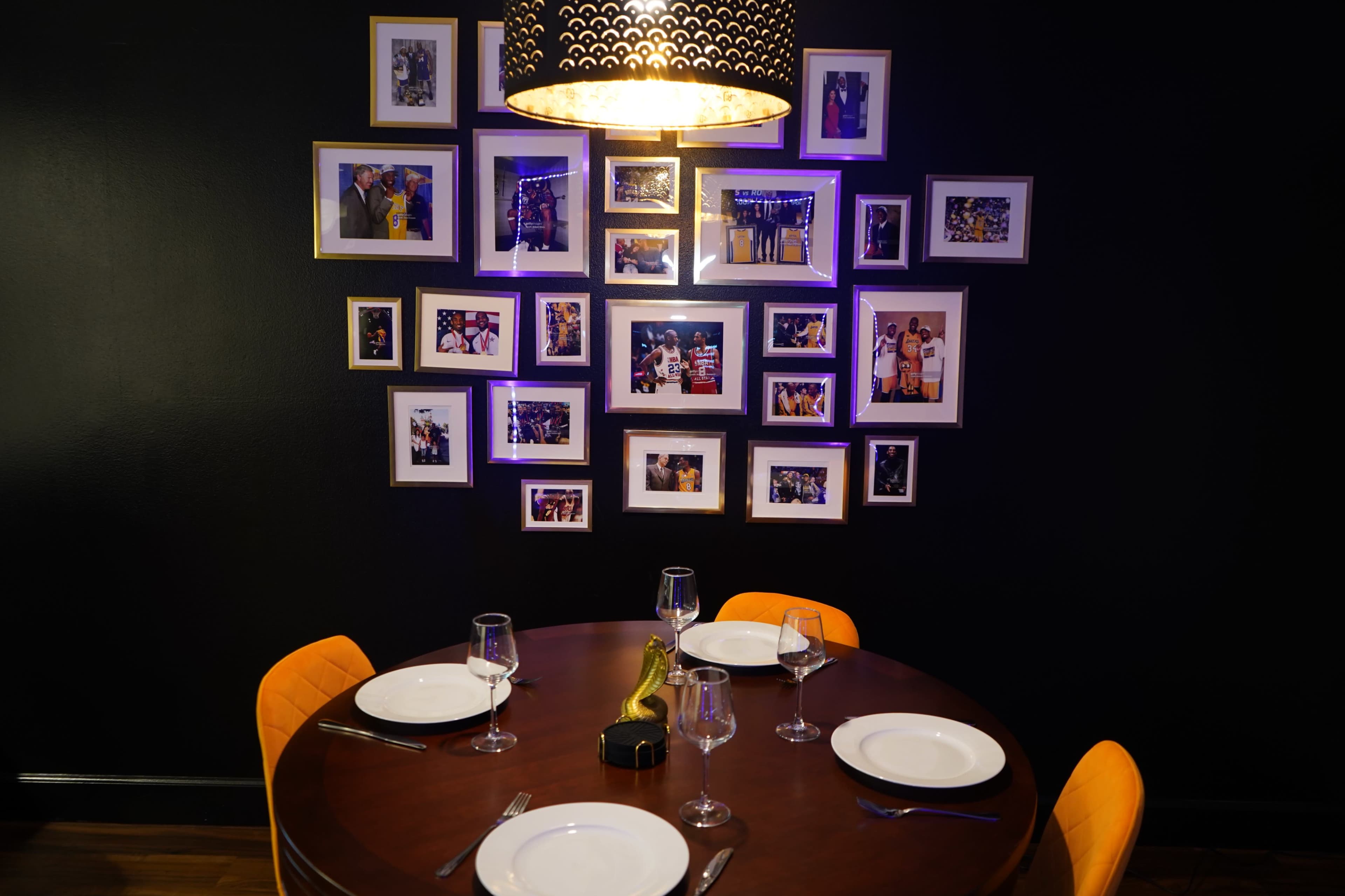A round dining table is set with plates and glasses, and a wall is decorated with framed photographs and a pendant light above.