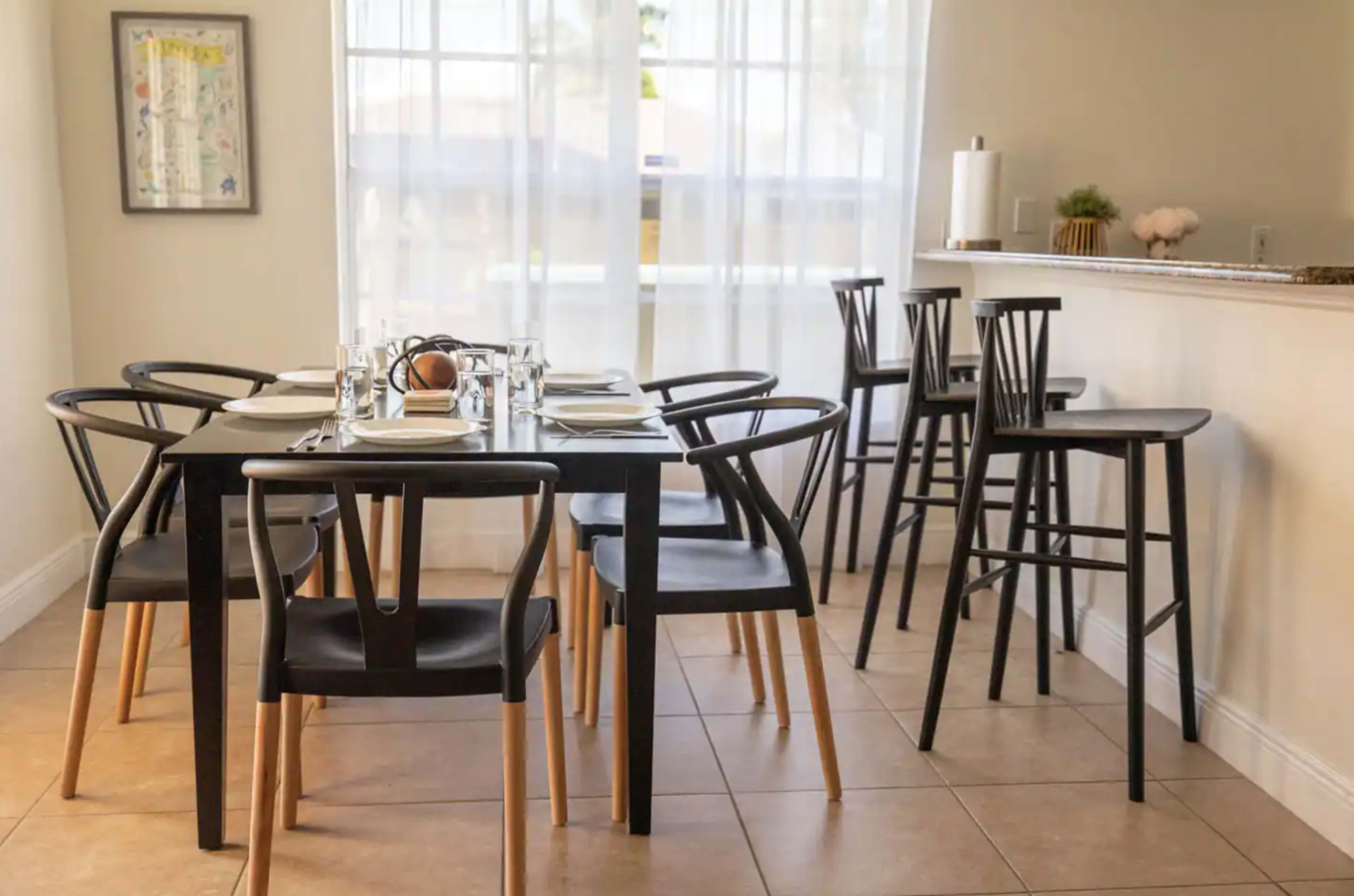 A dining area features a rectangular table set for a meal surrounded by black chairs, with high stools positioned along a nearby counter.