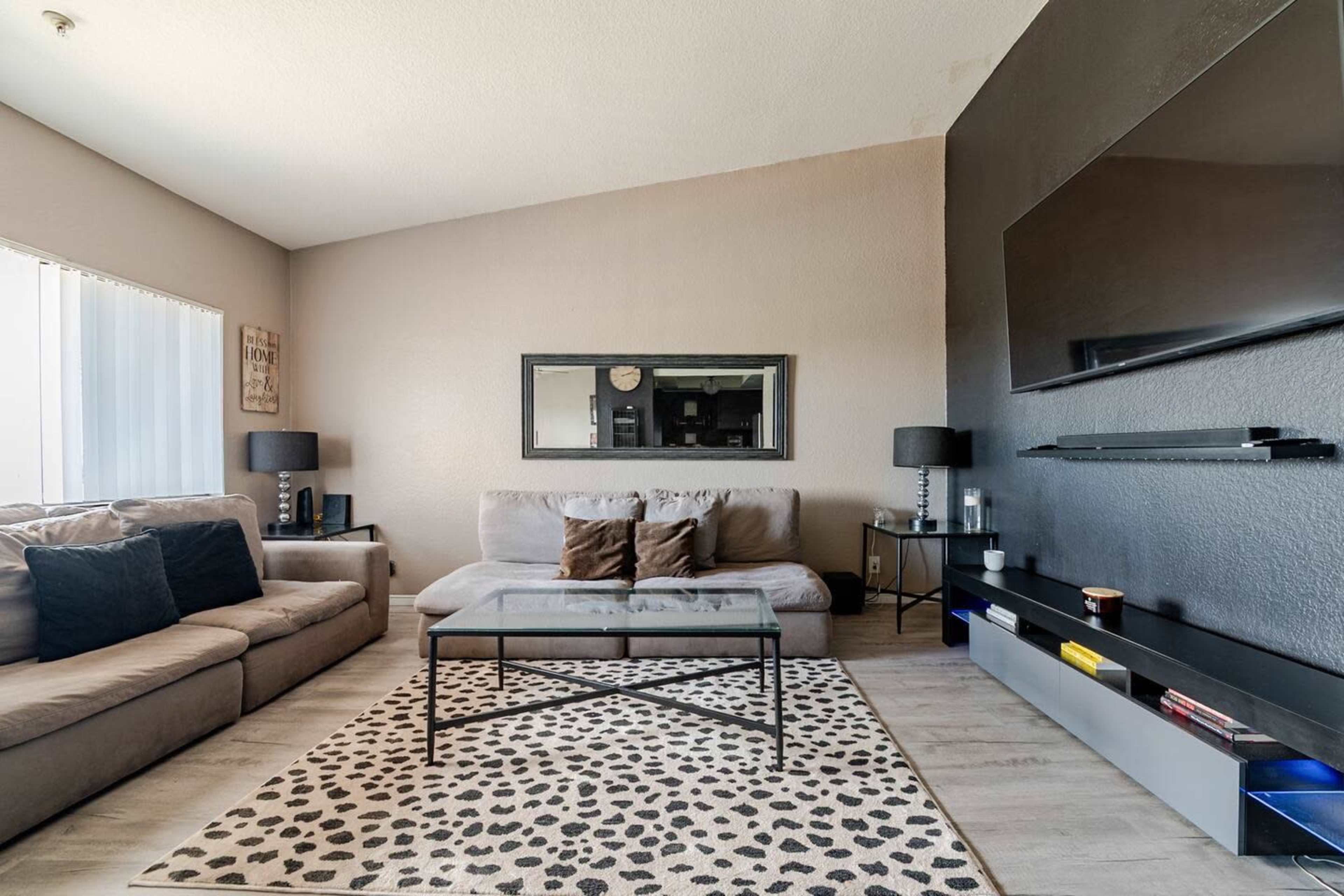 The room features a modern living area with a beige sectional sofa, a glass coffee table on a patterned rug, and a wall-mounted television.