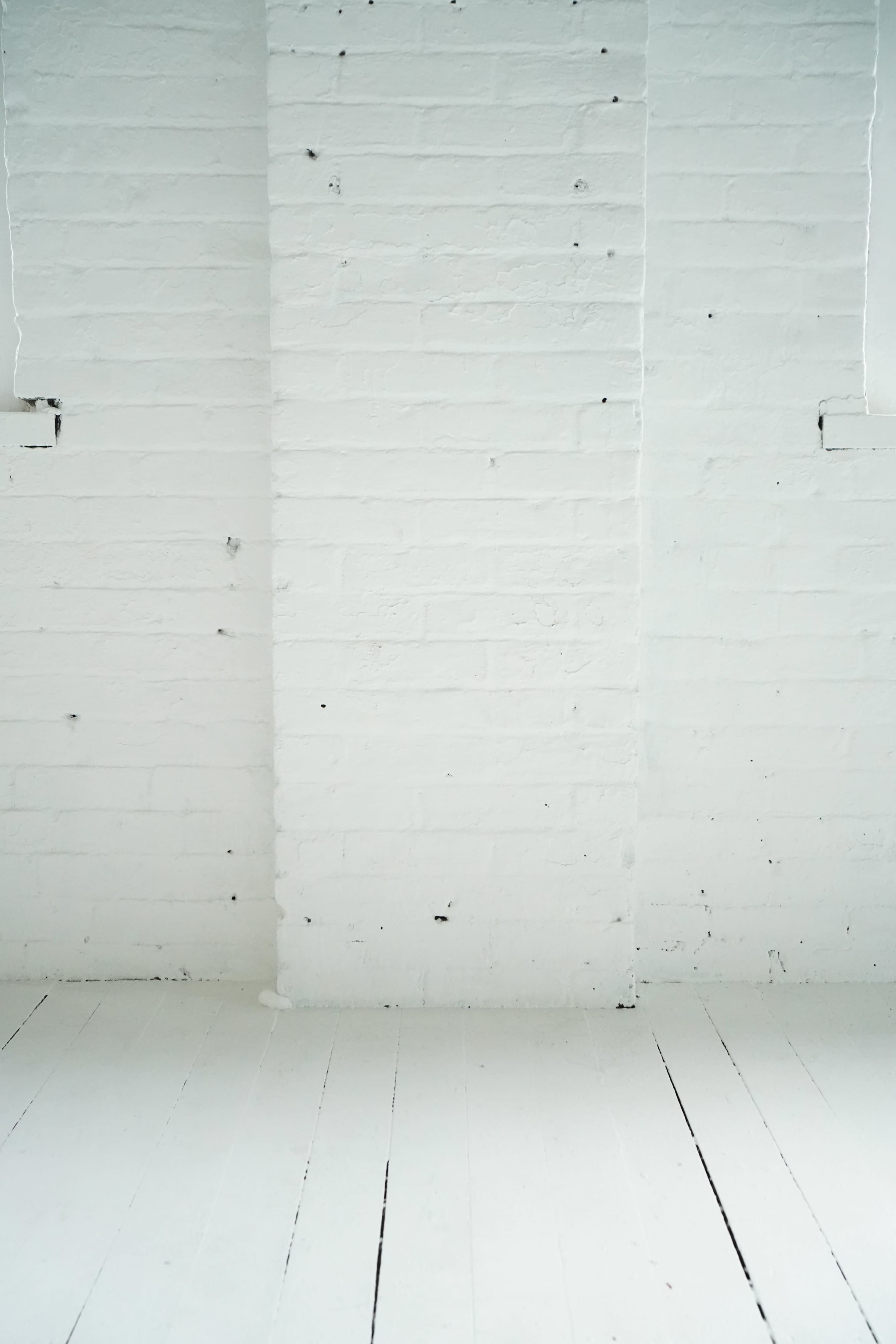 The image shows a minimalistic interior featuring a plain white brick wall and a light wooden floor.