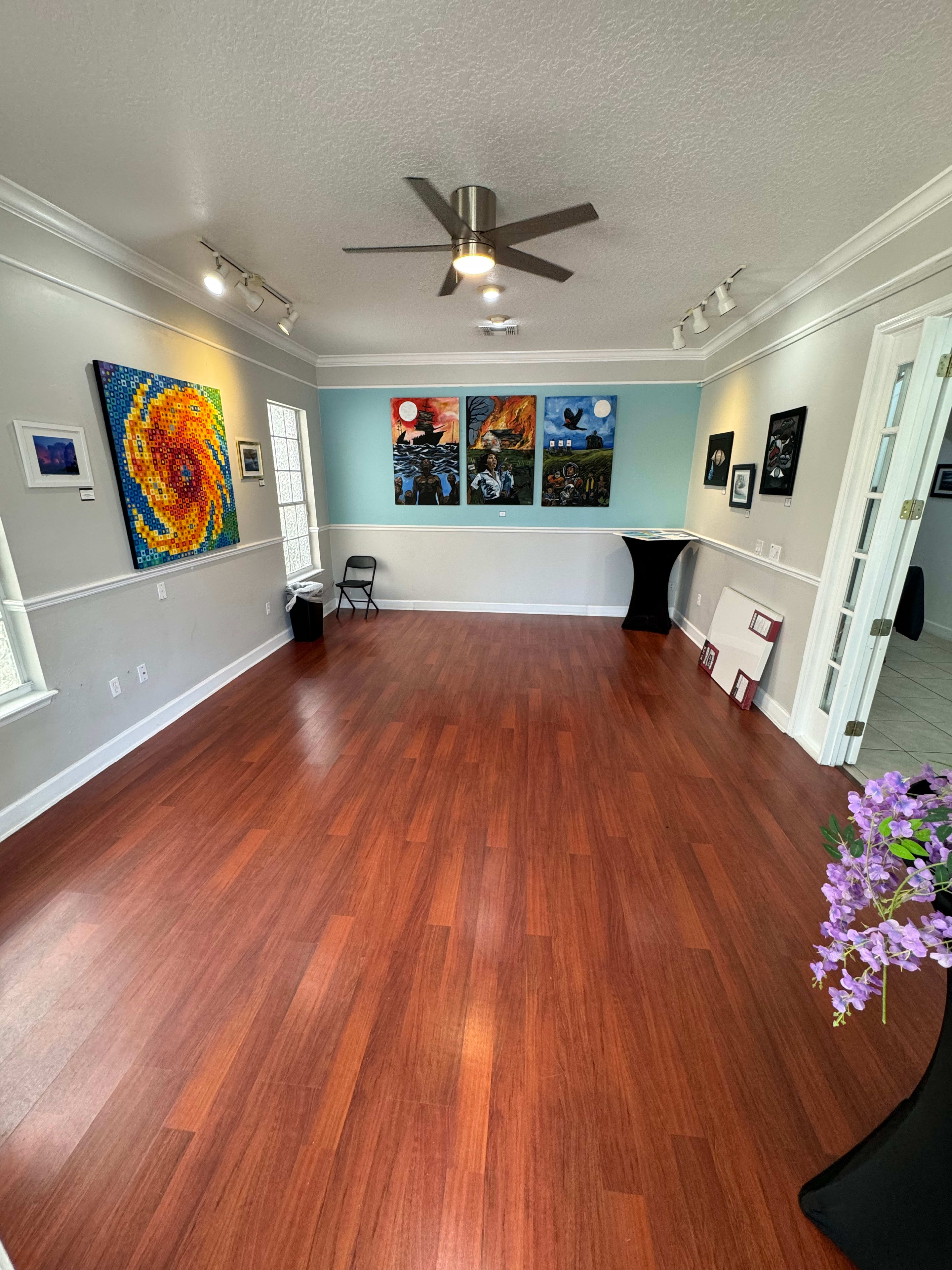 The image shows a brightly lit art gallery with wooden flooring, featuring colorful paintings on the walls and a small table in the corner.