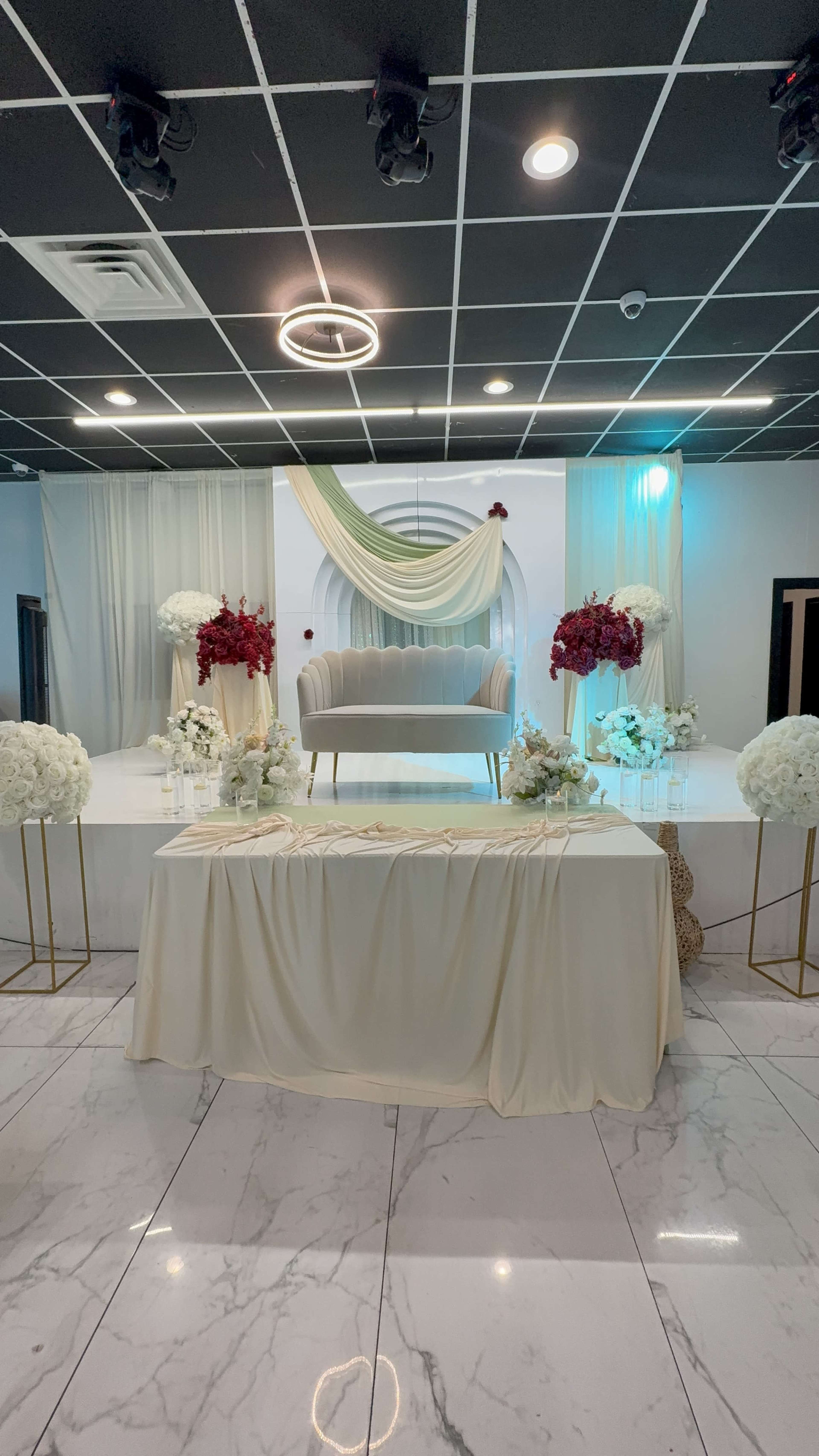 The image shows an elegantly decorated event space with a sofa on a raised platform, surrounded by floral arrangements and a table draped in fabric.