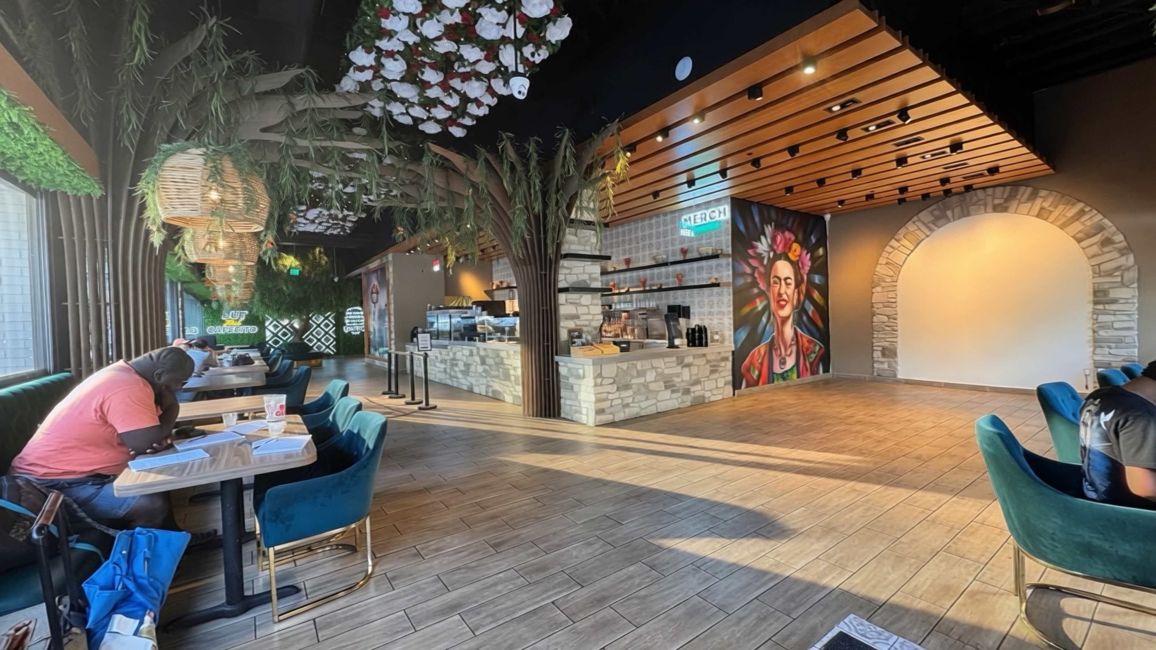 The image shows a modern café interior with lush greenery, a colorful mural of Frida Kahlo, and a spacious seating area featuring blue chairs and wooden tables.