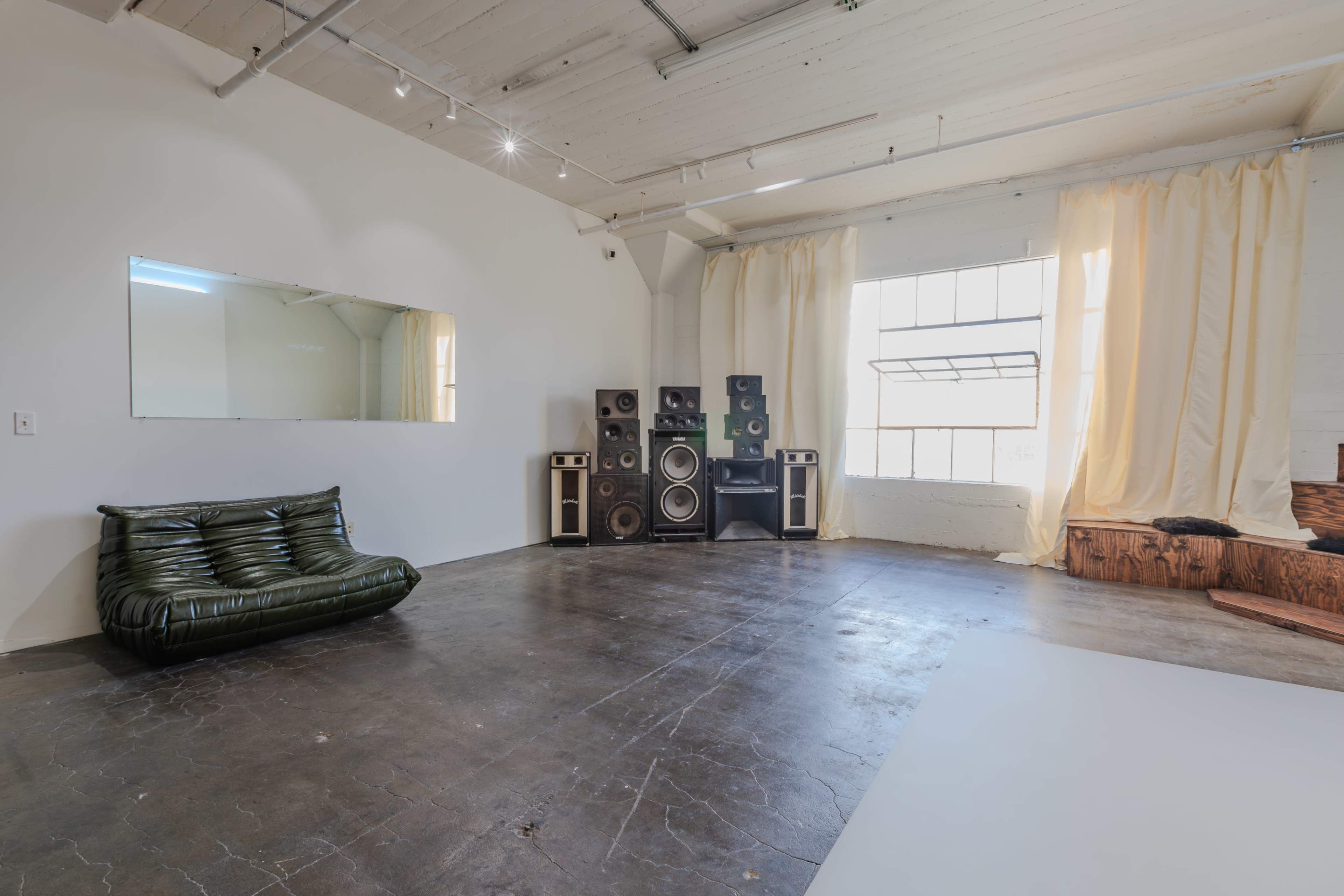 The image shows a spacious room with a green sofa, large speakers against the wall, and a window dressed with sheer curtains.