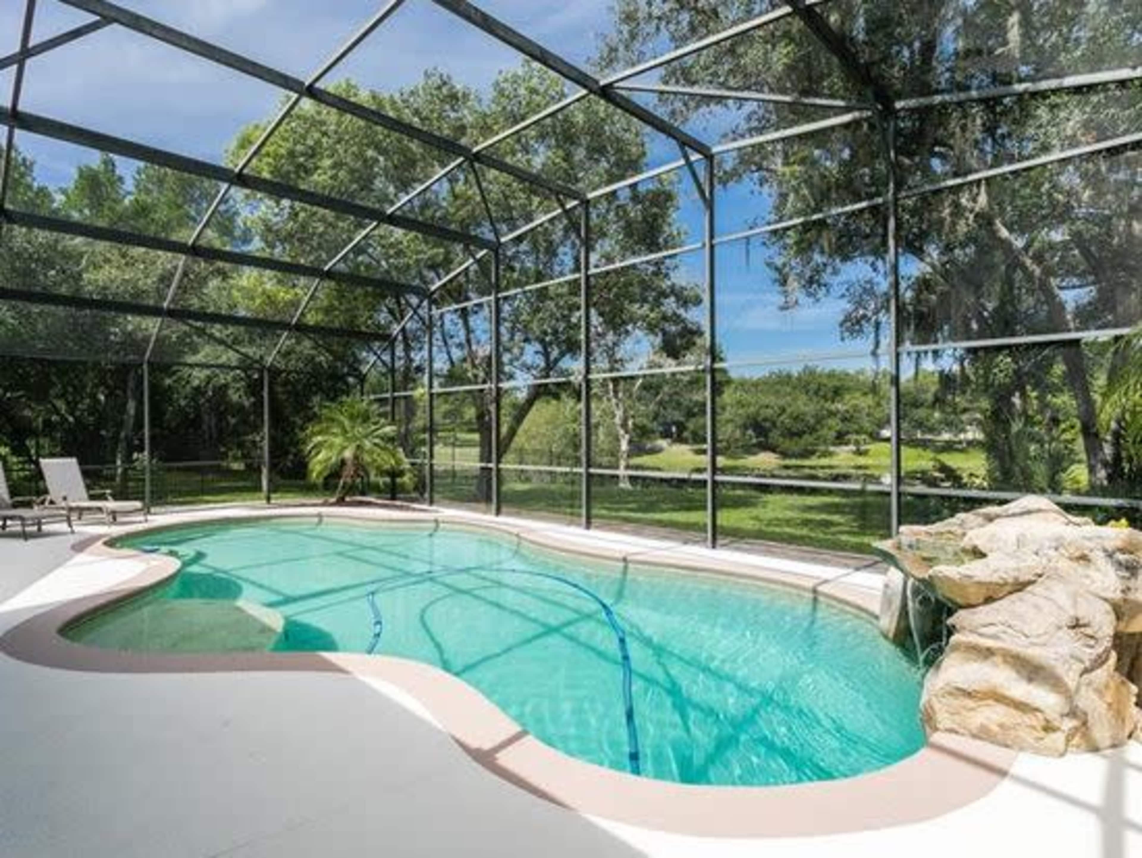 A screened-in pool area with a curved, turquoise pool surrounded by greenery and a rock feature.