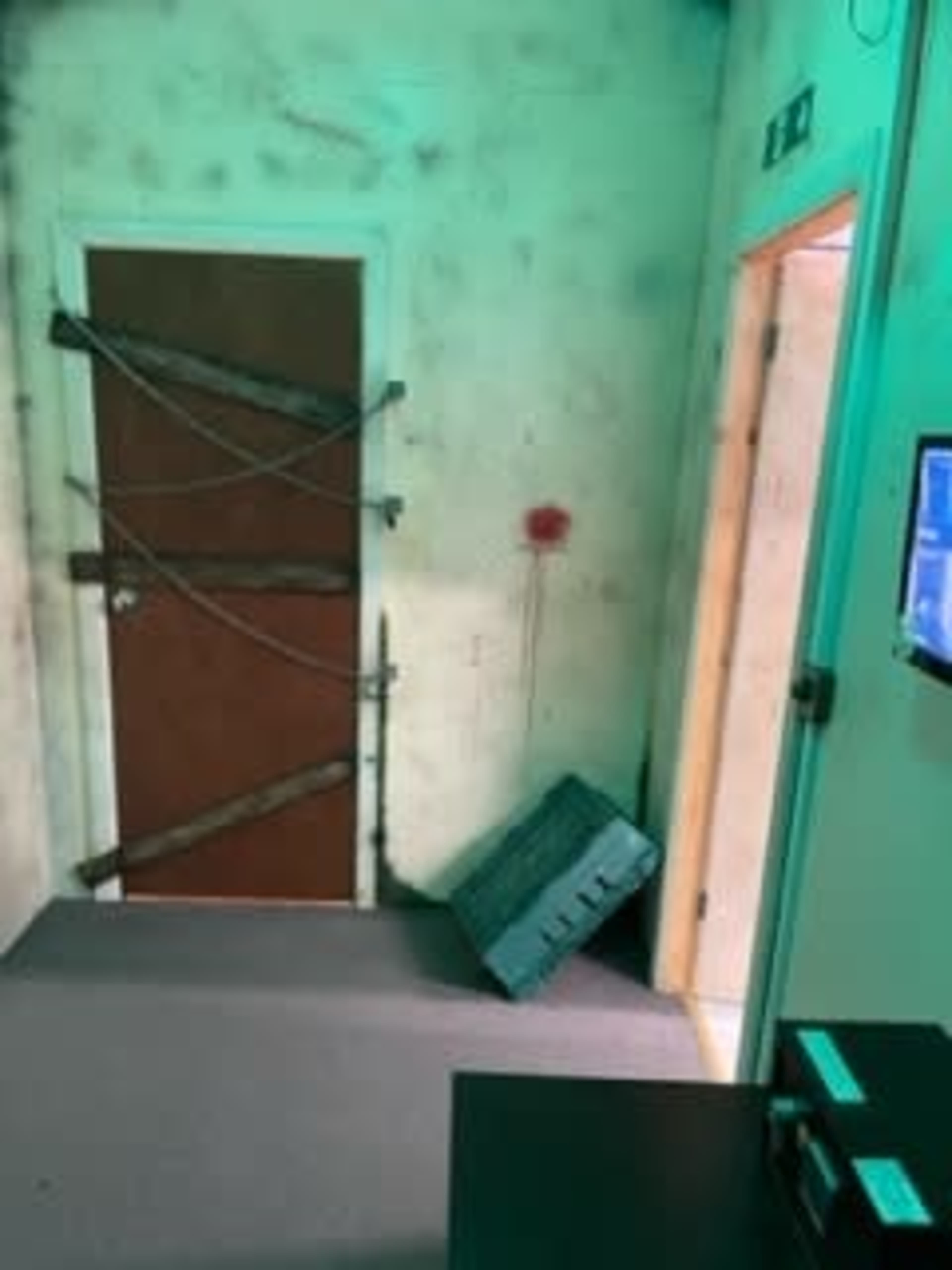 A dimly lit room with a door reinforced by wooden planks, a bloody mark on the wall, and a tilted crate on the floor.