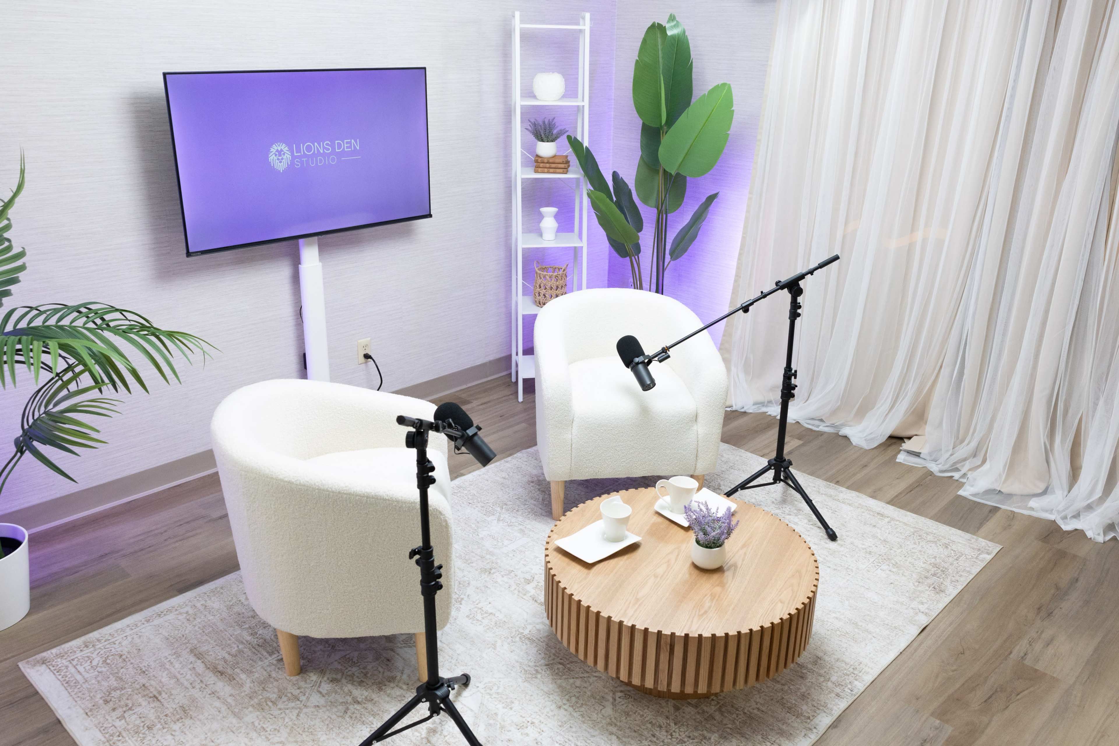 The image shows a modern studio setup featuring two white armchairs, a wooden coffee table with decorative items, and a television displaying the "Lions Den Studio" logo, surrounded by plants and soft drapery.