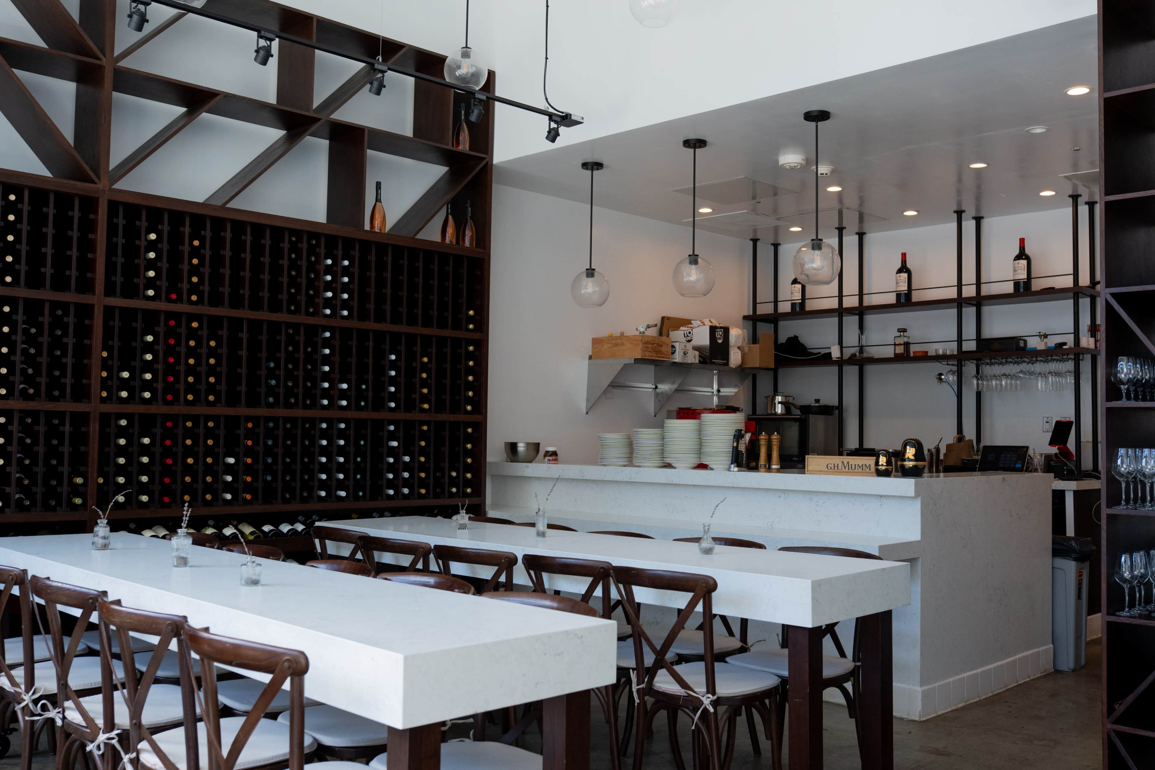 The image shows a modern restaurant interior featuring a long white table with wooden chairs, a wine storage wall, and an open kitchen area with pendant lighting.