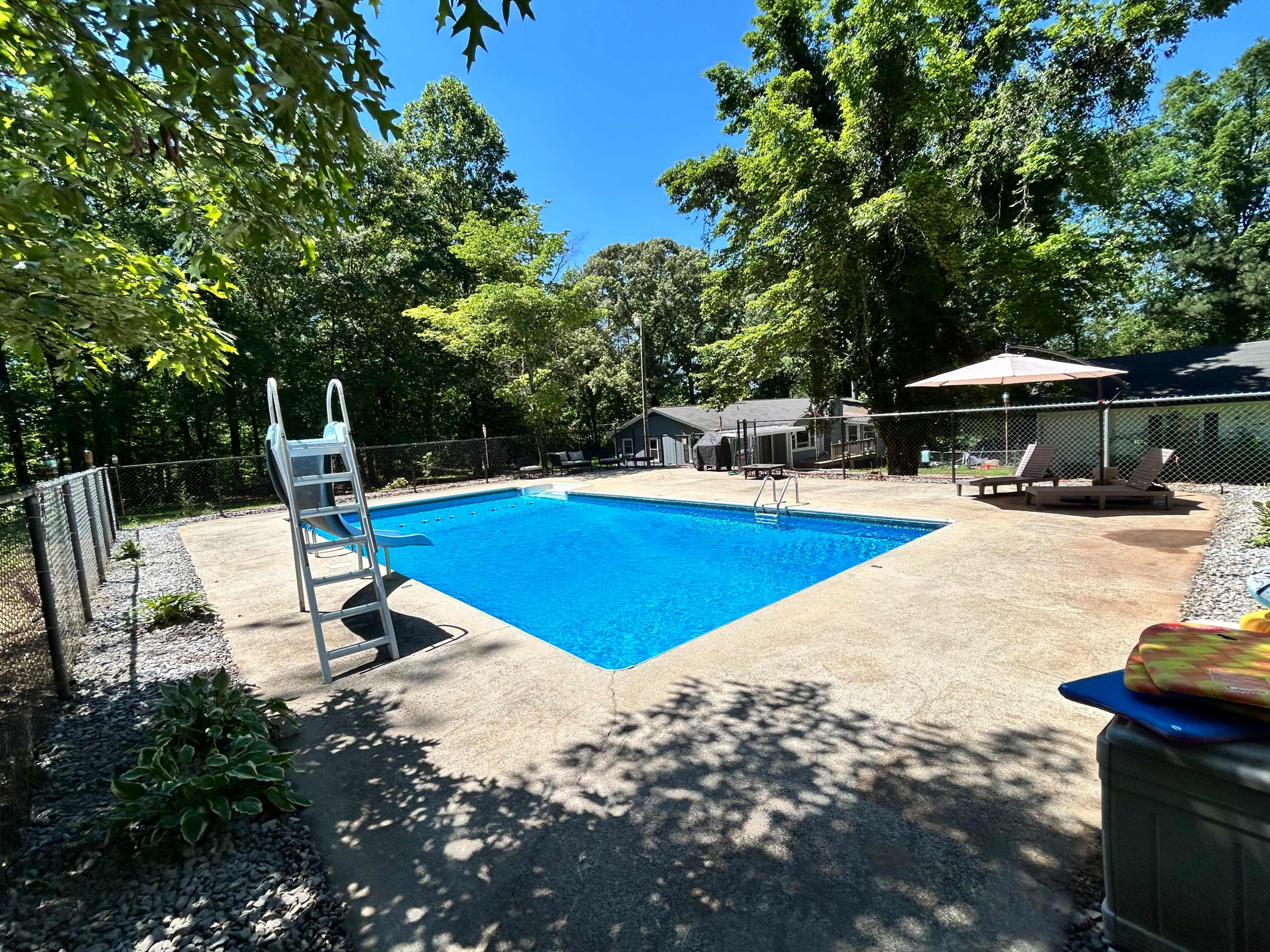 A fenced outdoor swimming pool surrounded by trees, with a diving board on one side and lounge chairs on the other.