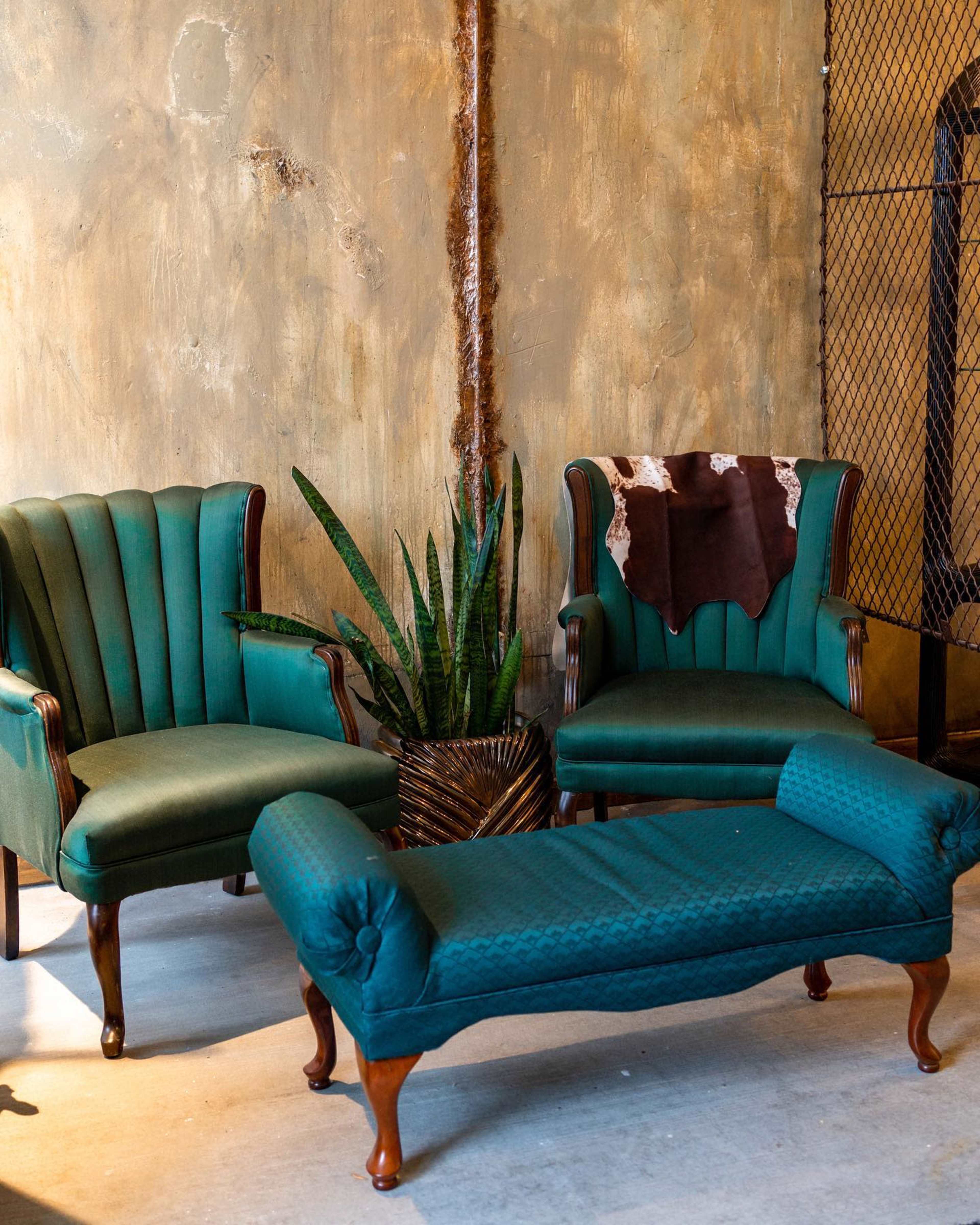 Two green upholstered armchairs and a tufted upholstered bench with wooden legs, placed against a textured wall and accompanied by a potted plant.