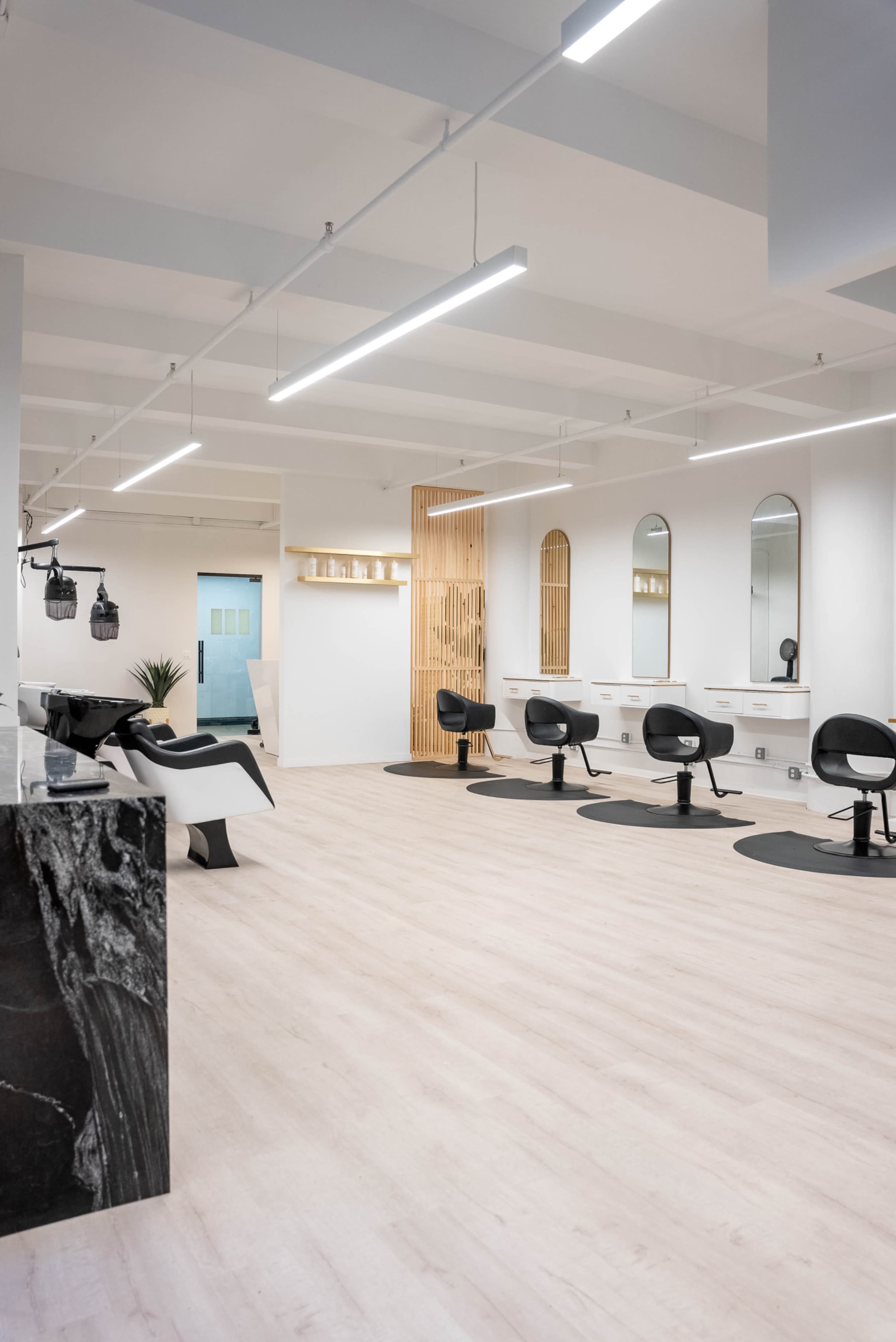 A modern hair salon with several styling stations, mirrors, and black salon chairs arranged on light-colored wooden flooring.