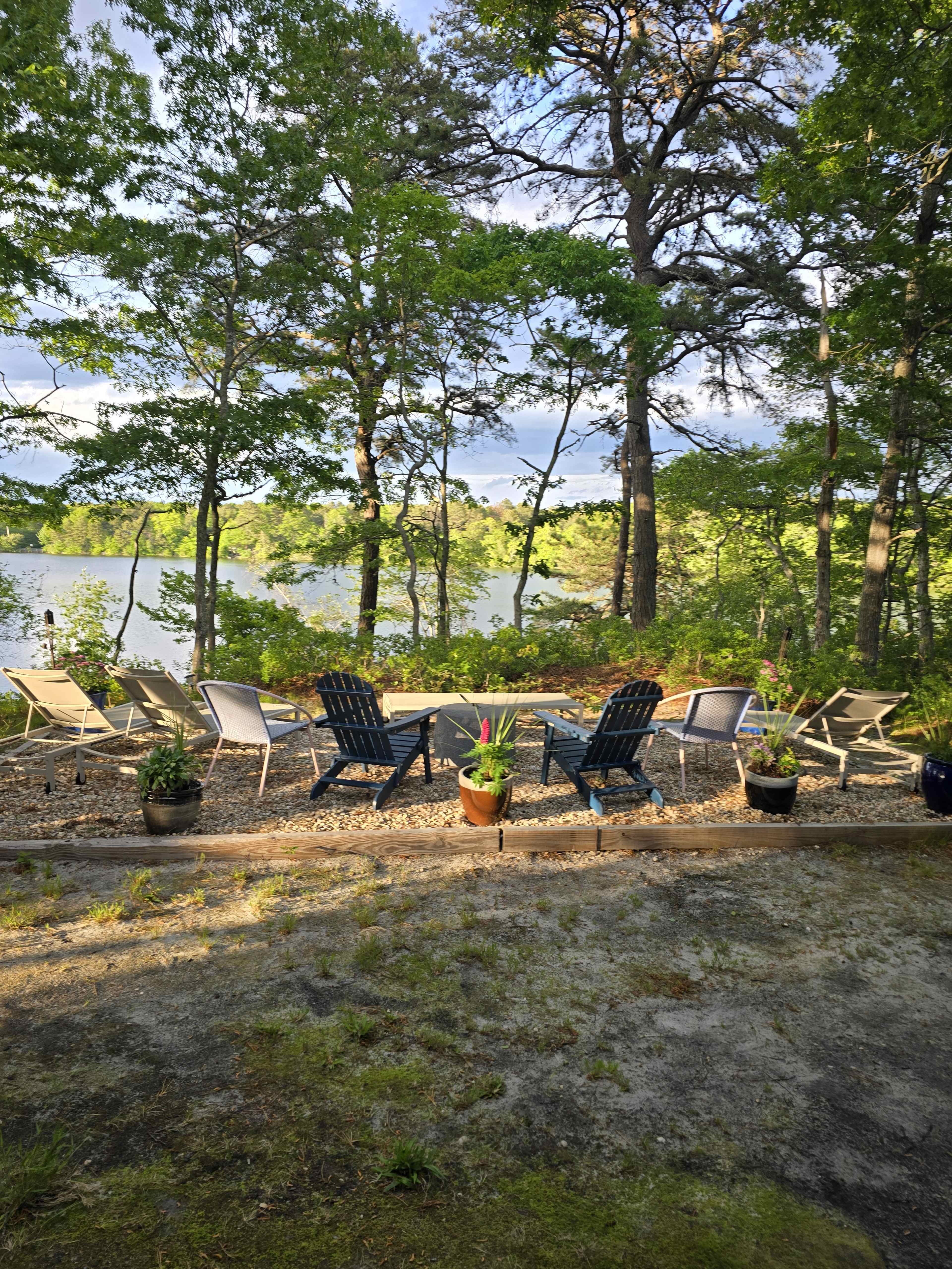 The image shows a serene outdoor setting with several lounge chairs arranged in a circle near a water body, surrounded by trees and potted plants.