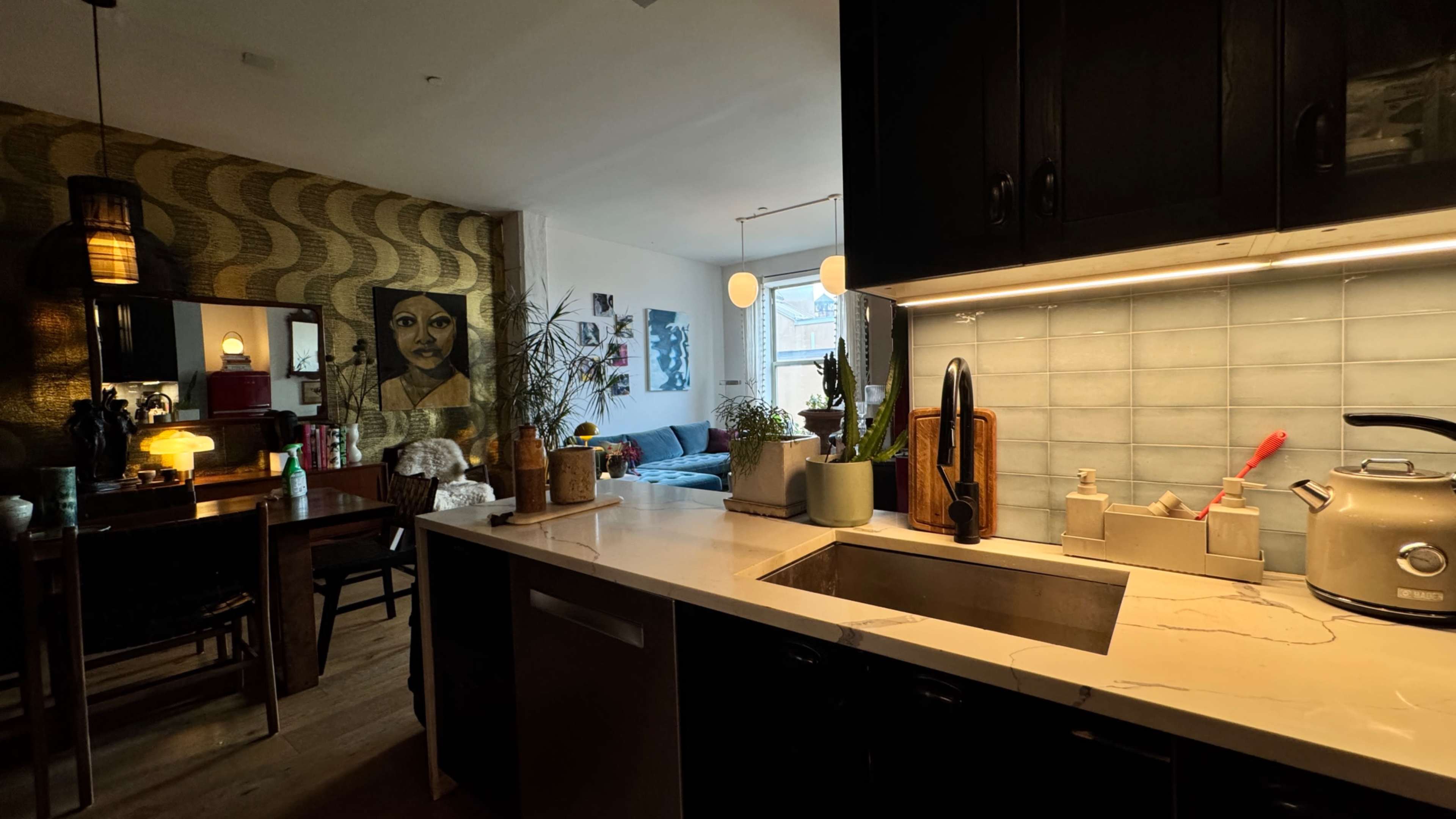 A modern kitchen features a sink and counter, with a dining area visible in the background, alongside a decorated wall and a plush sofa.