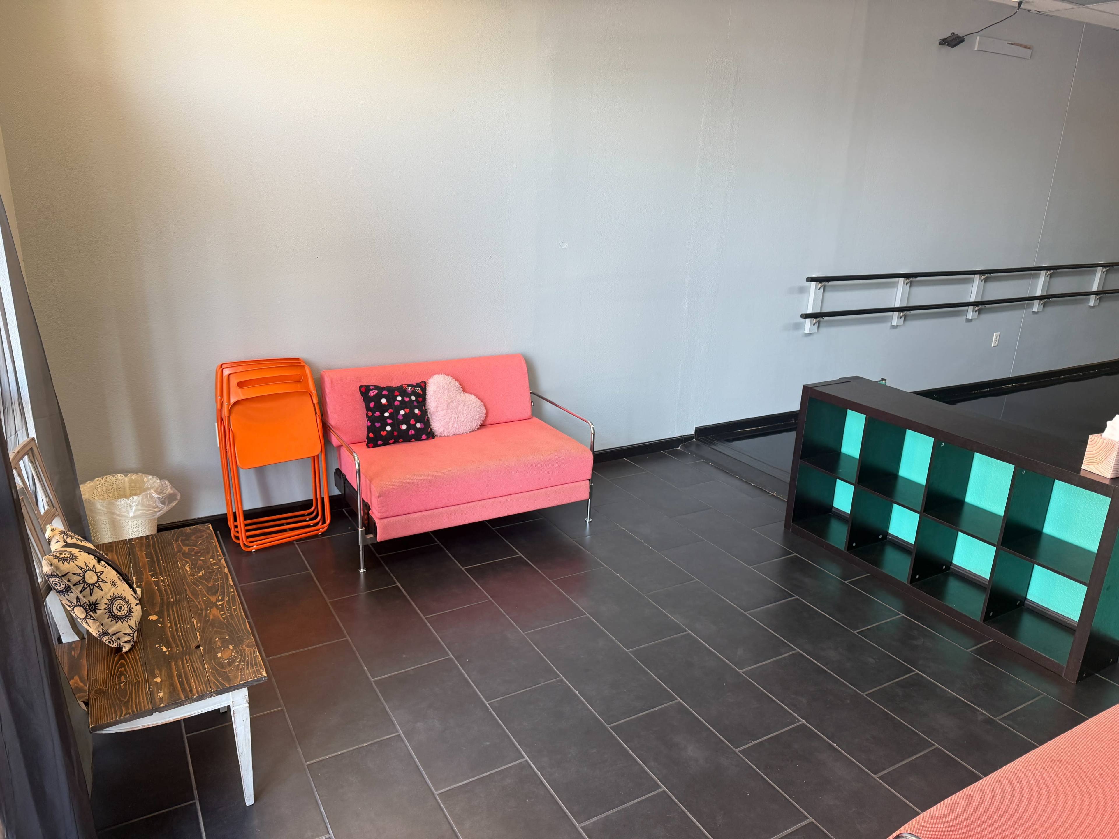 The image shows a spacious room with a pink couch, stacked orange chairs, a black and teal shelving unit, and tiled flooring.