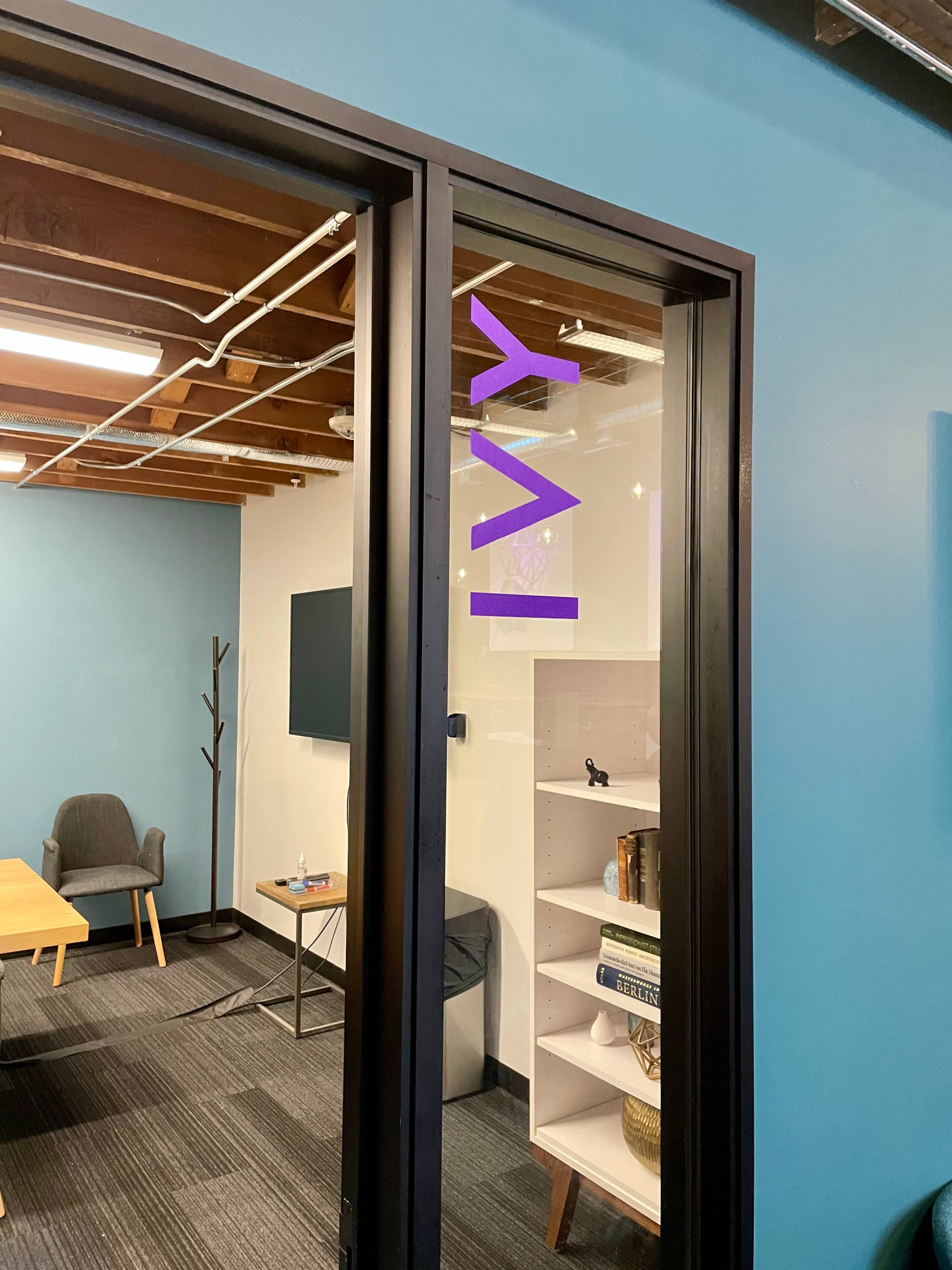 A glass door with the word "IVY" in purple lettering opens into an office space featuring a chair, a television, and a bookshelf.