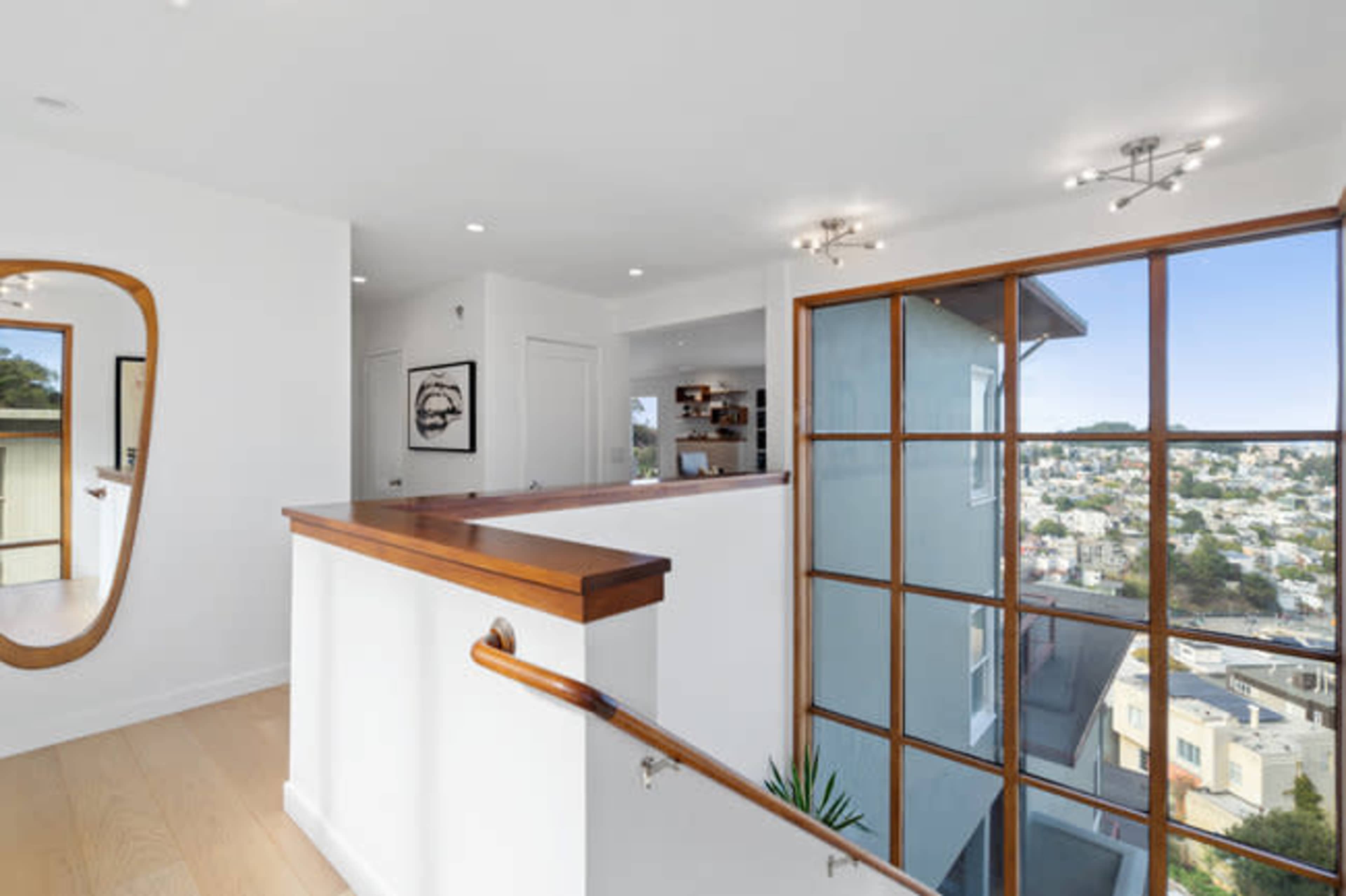 The image shows a modern hallway with a wooden railing, a large mirror on the wall, and a window displaying a view of a cityscape.