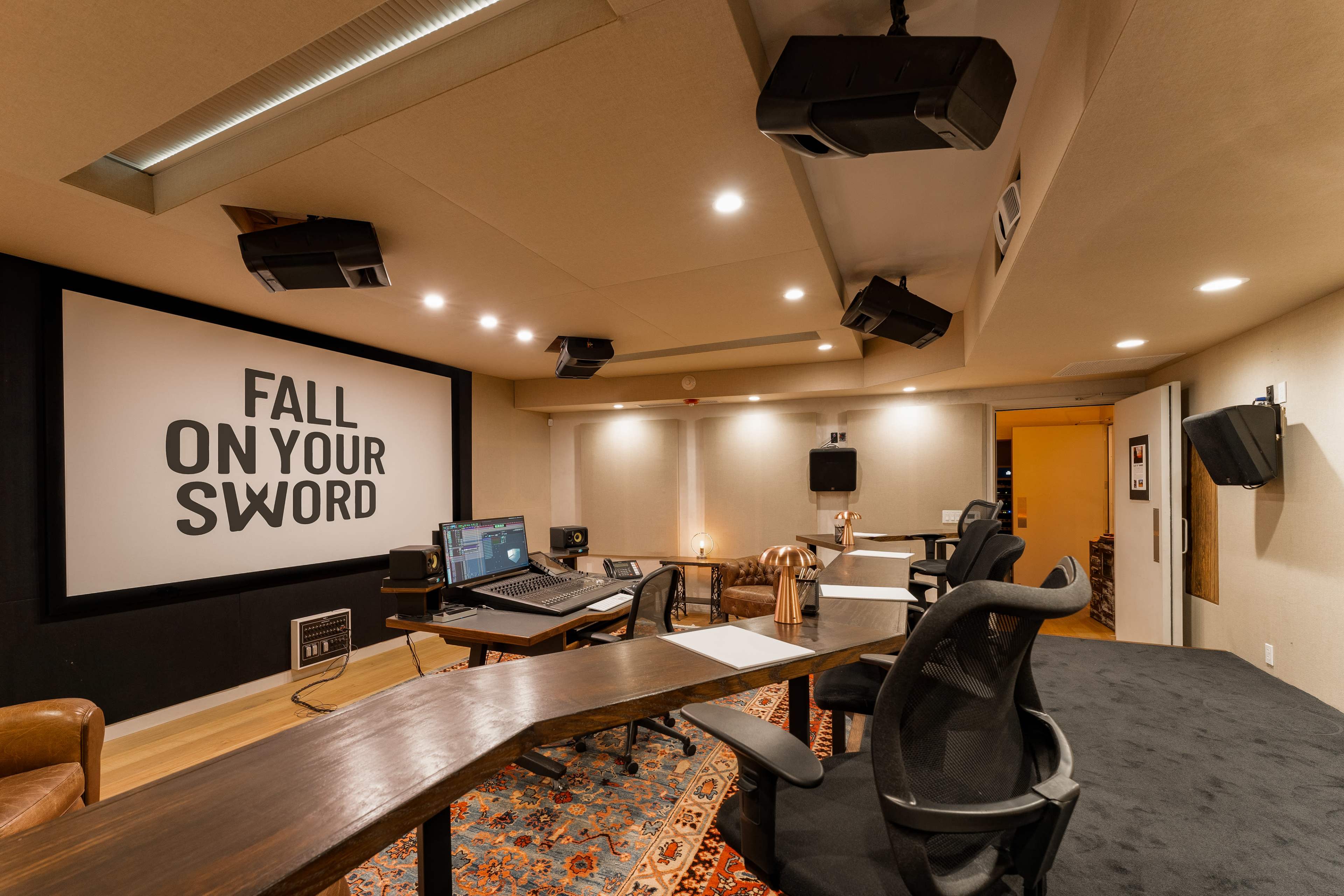 A modern recording studio with dark walls, a large screen displaying the phrase "FALL ON YOUR SWORD," and multiple audio equipment setups.