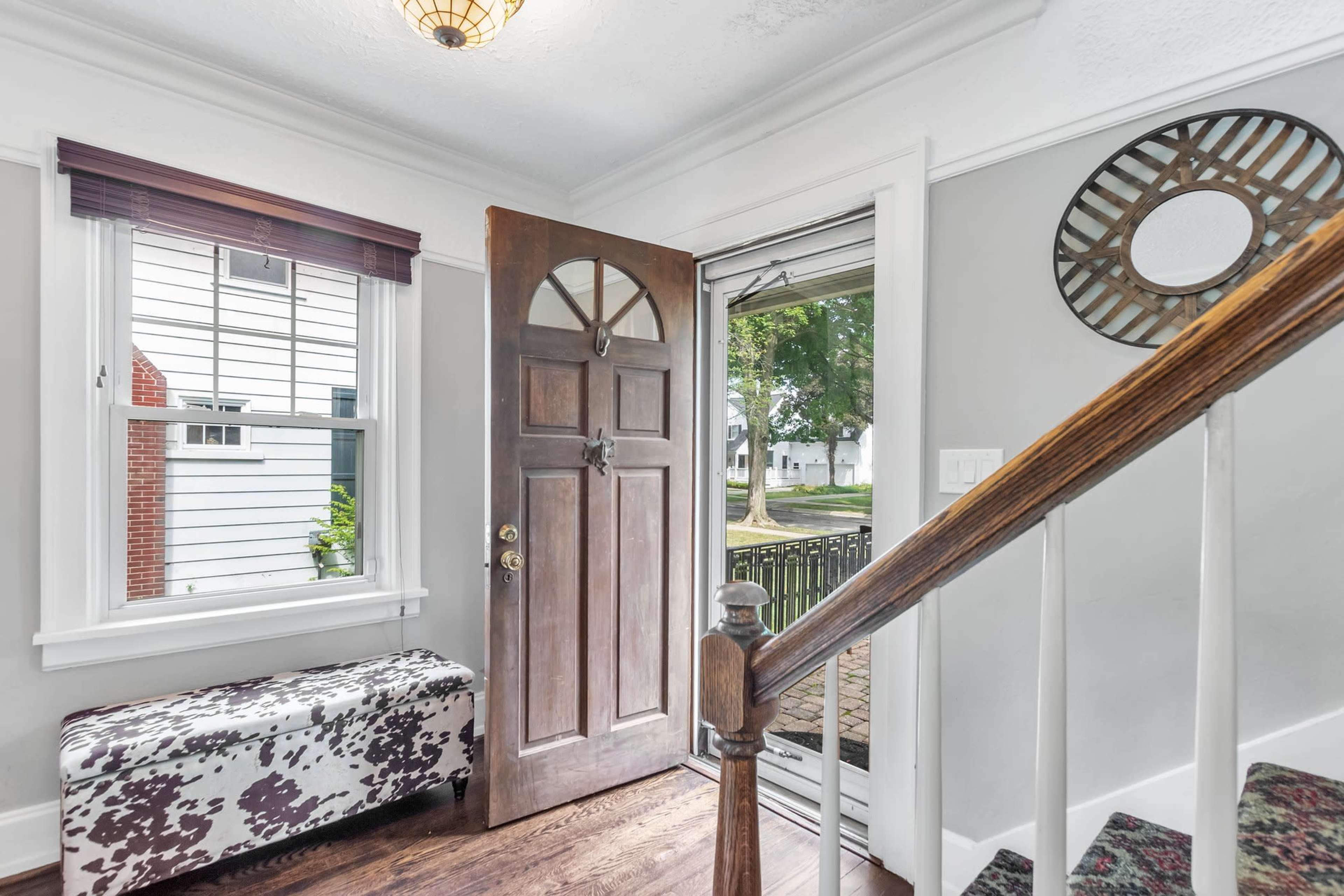 An entryway with a brown front door open to a view of a porch and exterior landscaping, along with a patterned bench and stairs leading to an upper level.