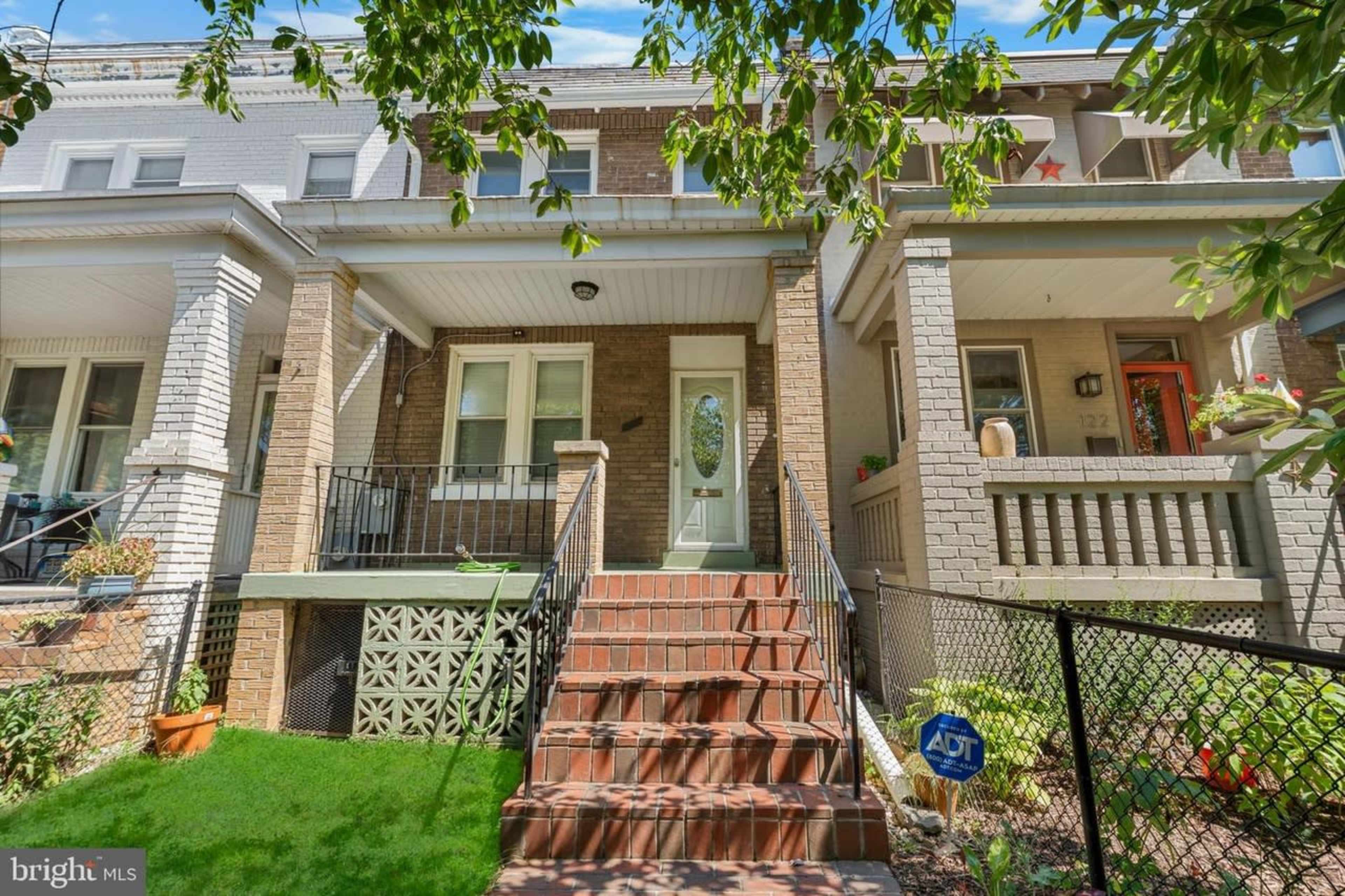 The image shows a row of three houses with brick facades, each featuring a front staircase and a porch, surrounded by greenery and a fence.