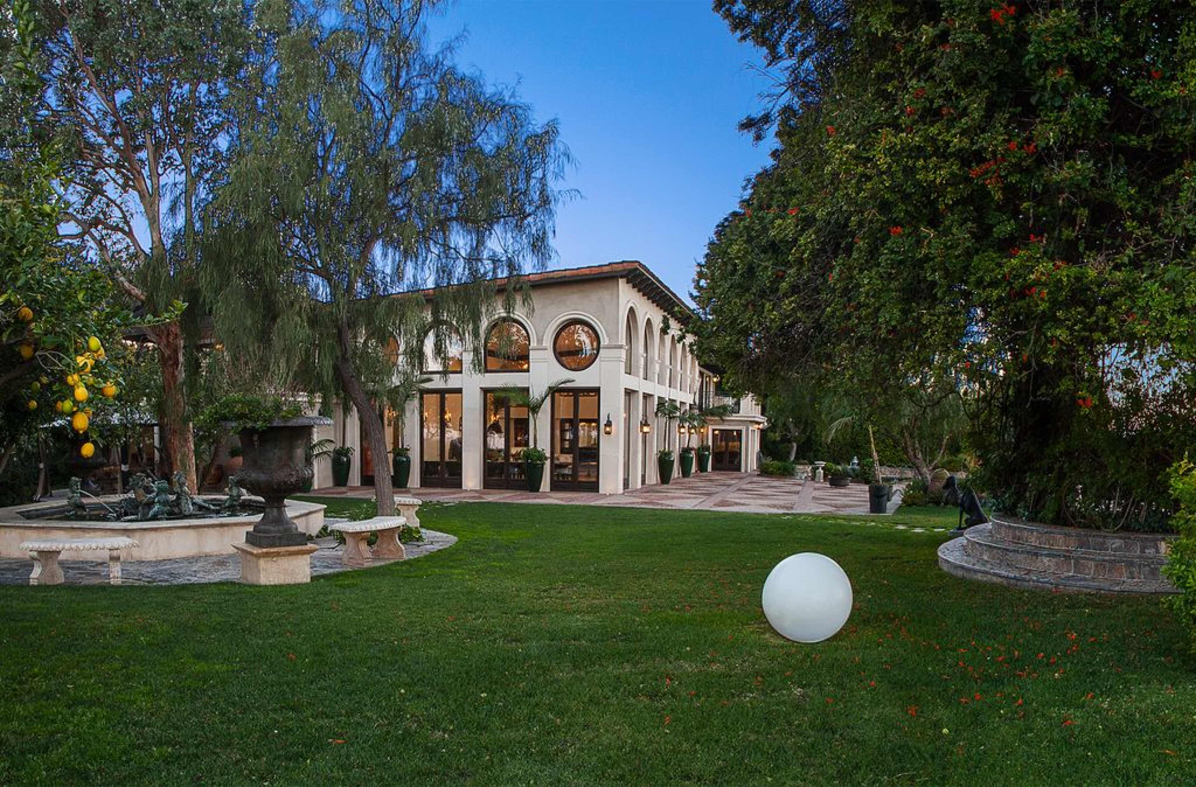 A large, two-story house with arched windows surrounded by a green lawn and trees, featuring a pool and decorative stone landscaping.