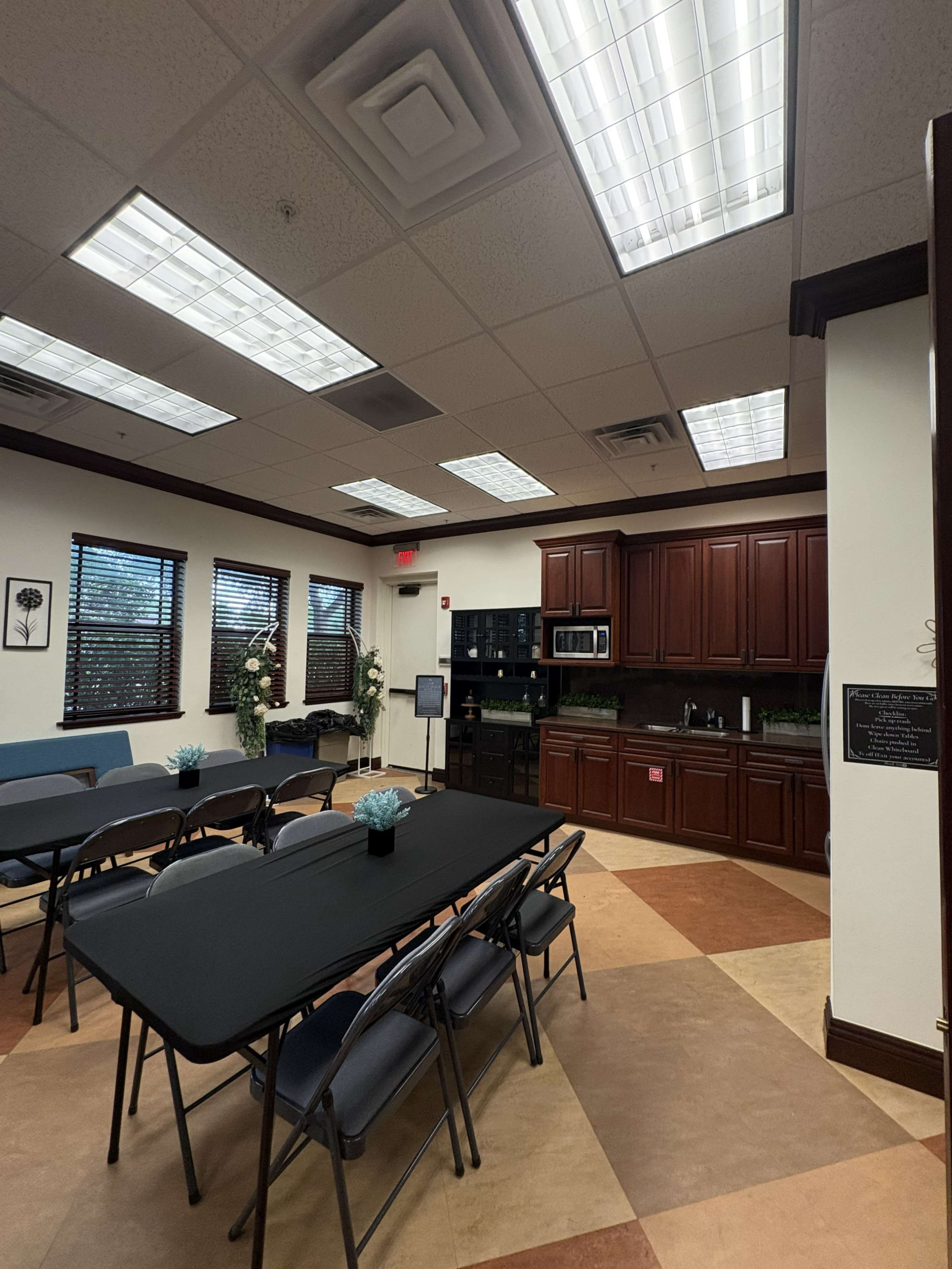 The image shows a meeting room with several black folding tables set up, a kitchenette area featuring cabinets and appliances, and large windows allowing natural light to enter.