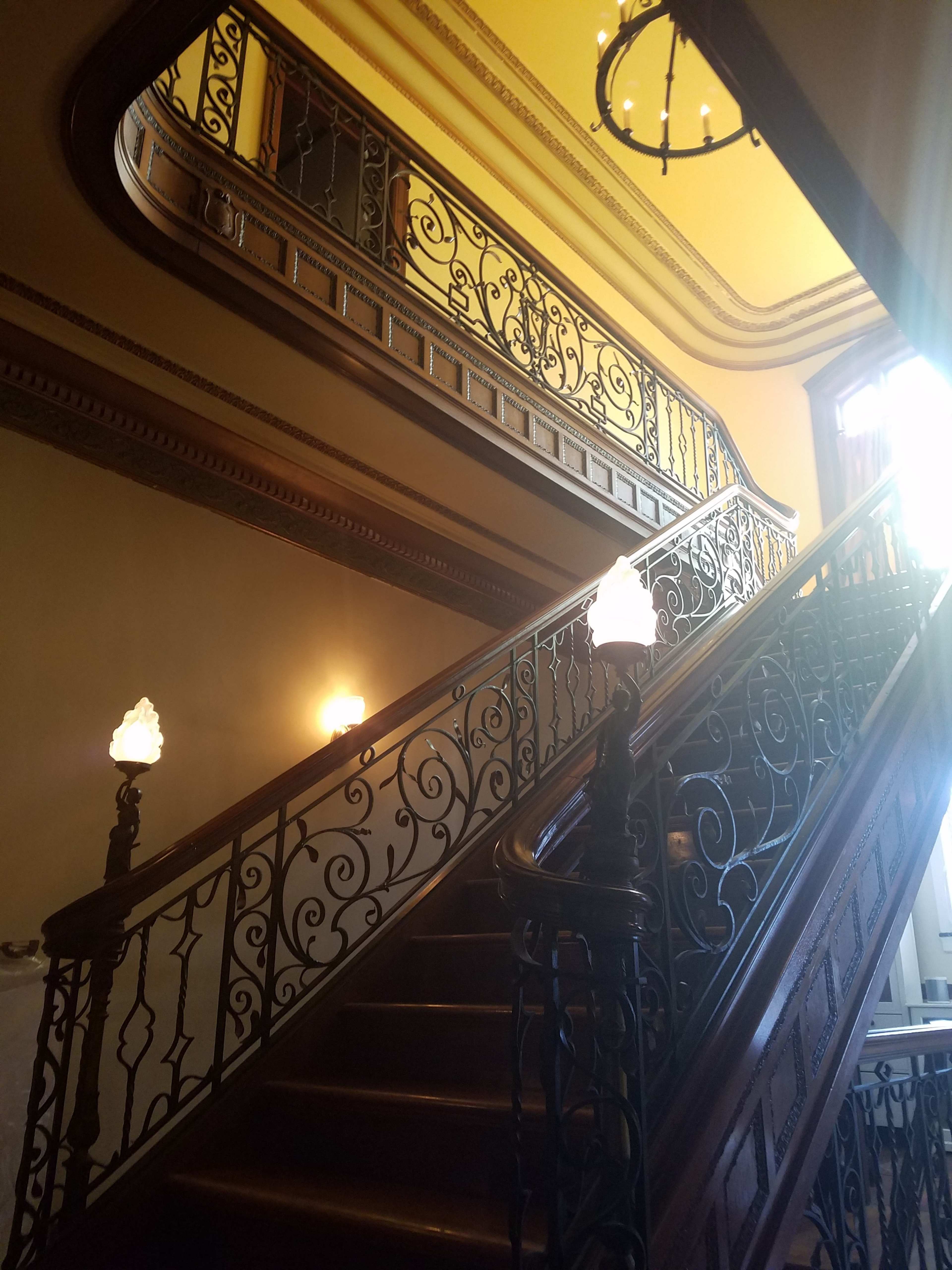 The image shows a grand staircase with intricate wrought iron railings, leading upward to a brightly lit upper level beneath a decorative ceiling.
