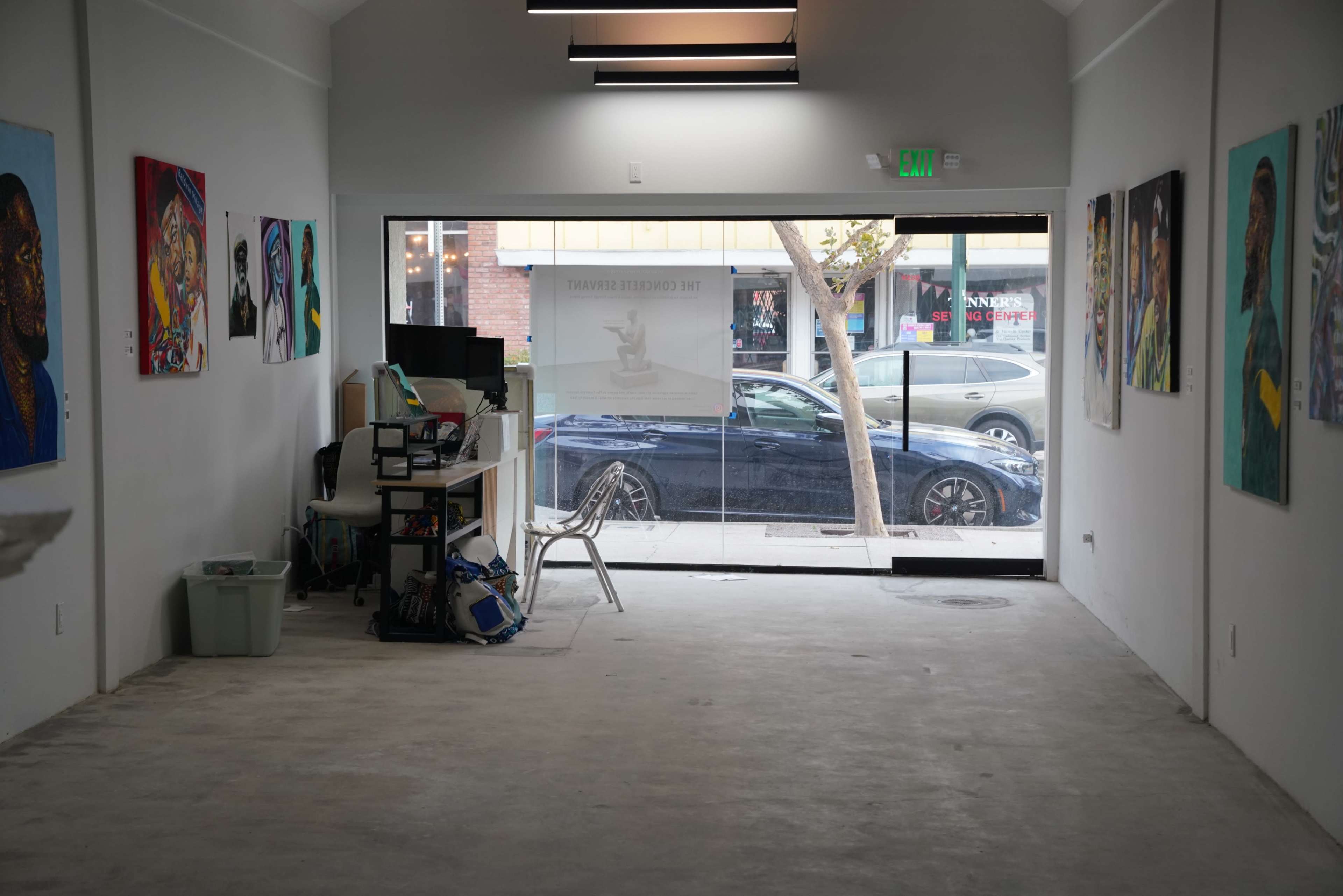 The image shows an empty art gallery with colorful paintings on the walls and a large window facing the street.