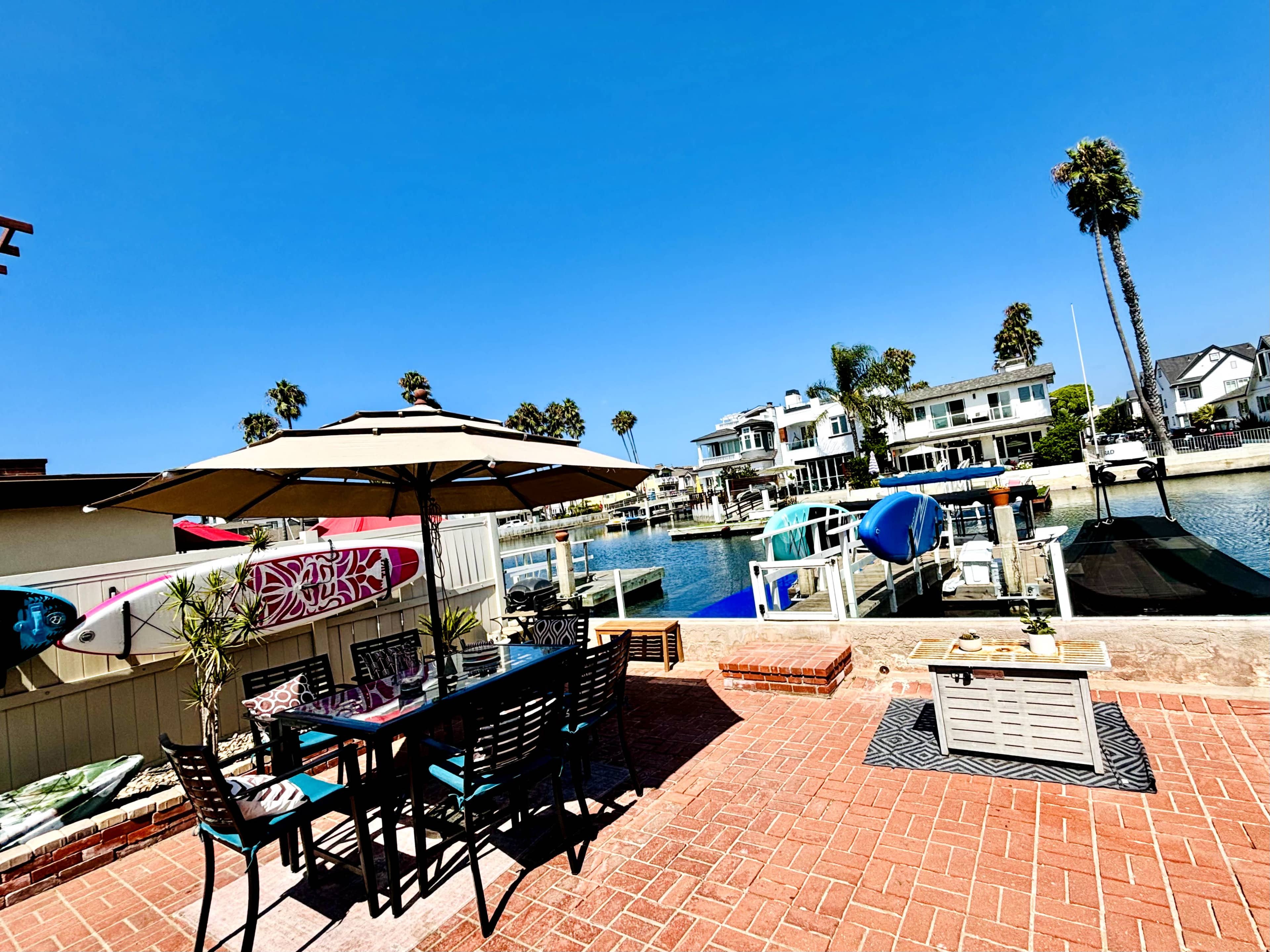 A patio with a dining table and umbrella overlooks a waterfront with boats and palm trees in the background.