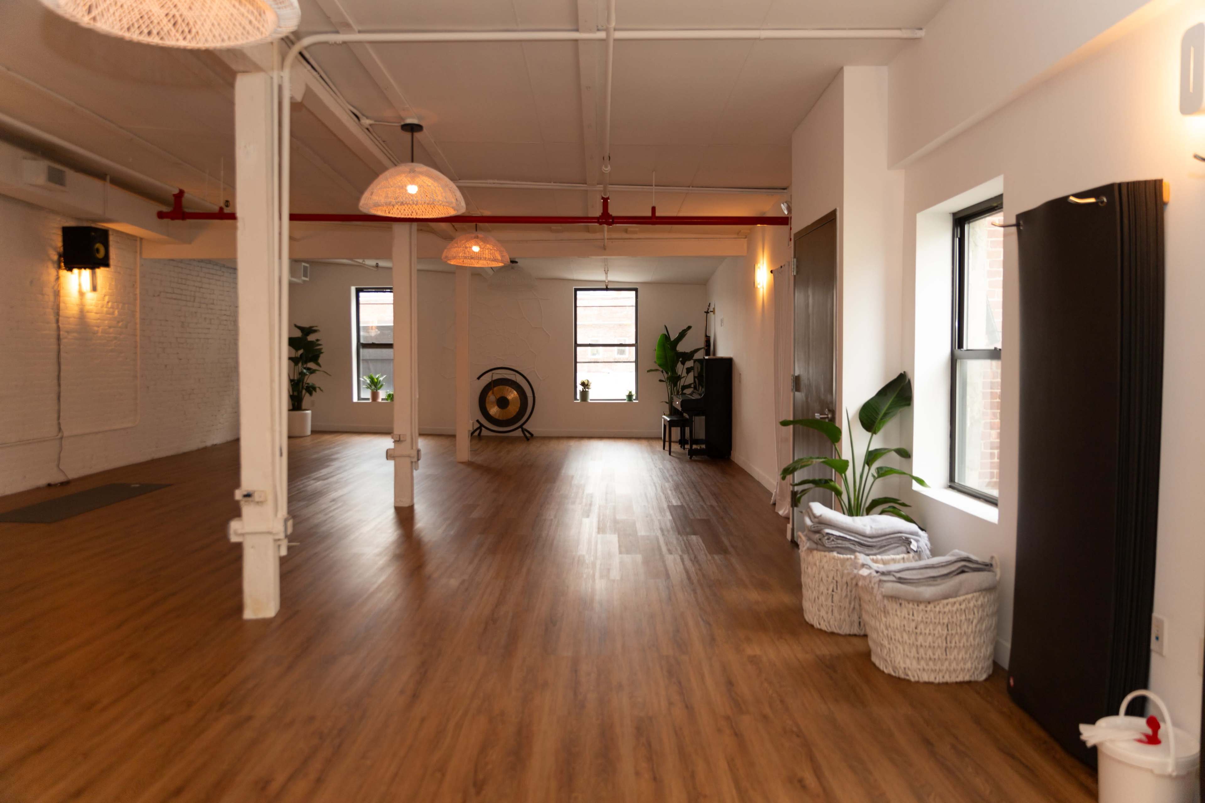 The image shows a spacious, well-lit studio with wooden flooring, natural light coming through windows, and plants in the corners.