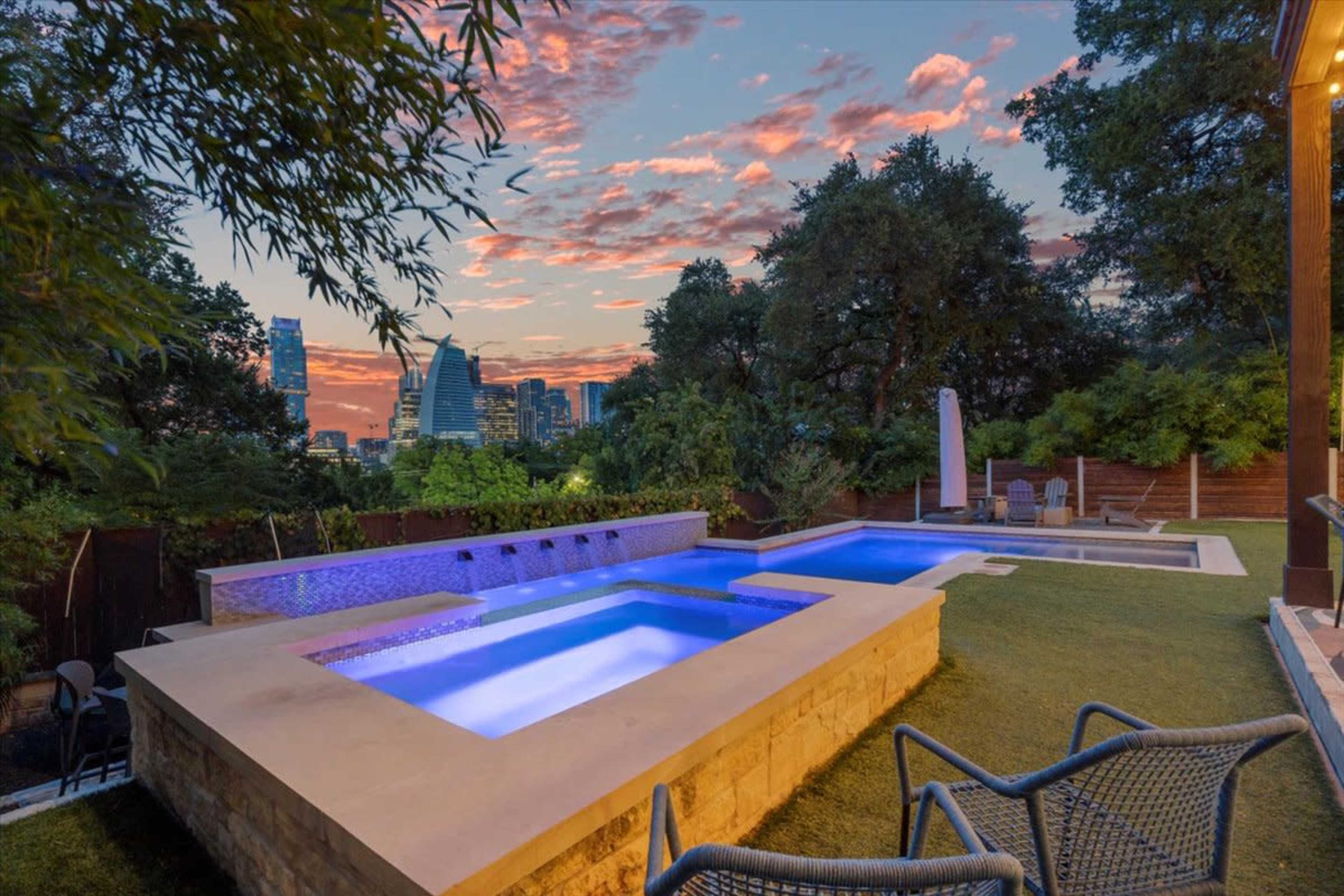 A modern backyard features a rectangular pool with a spa, surrounded by lush greenery and a city skyline at sunset.