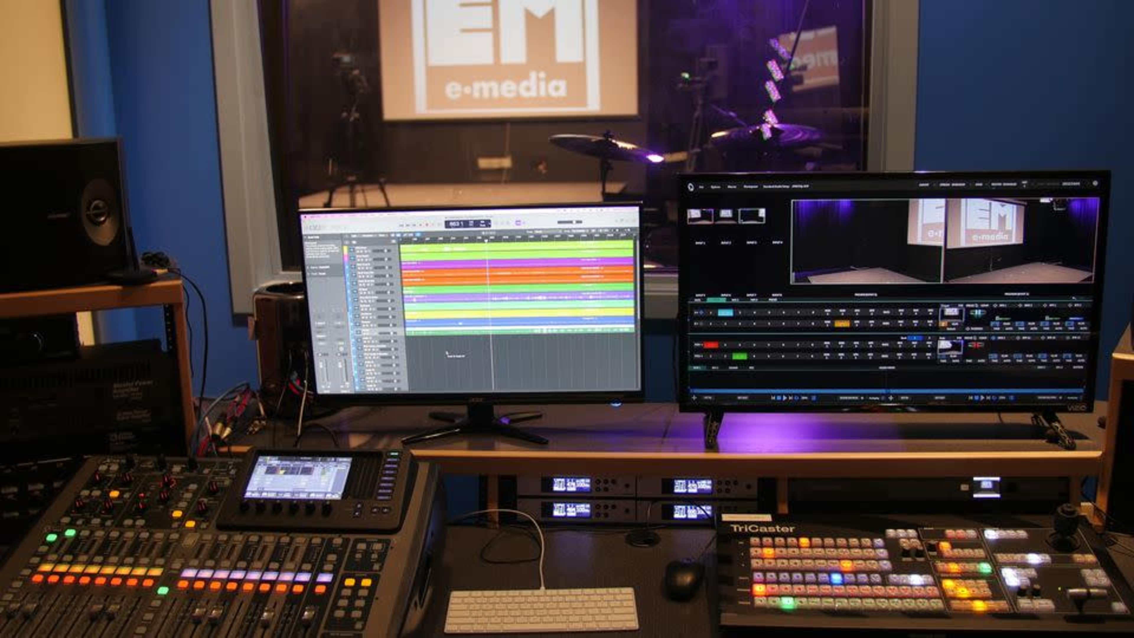 A professional media production setup with two large monitors displaying audio and video editing software, alongside a control panel for managing various media functions.