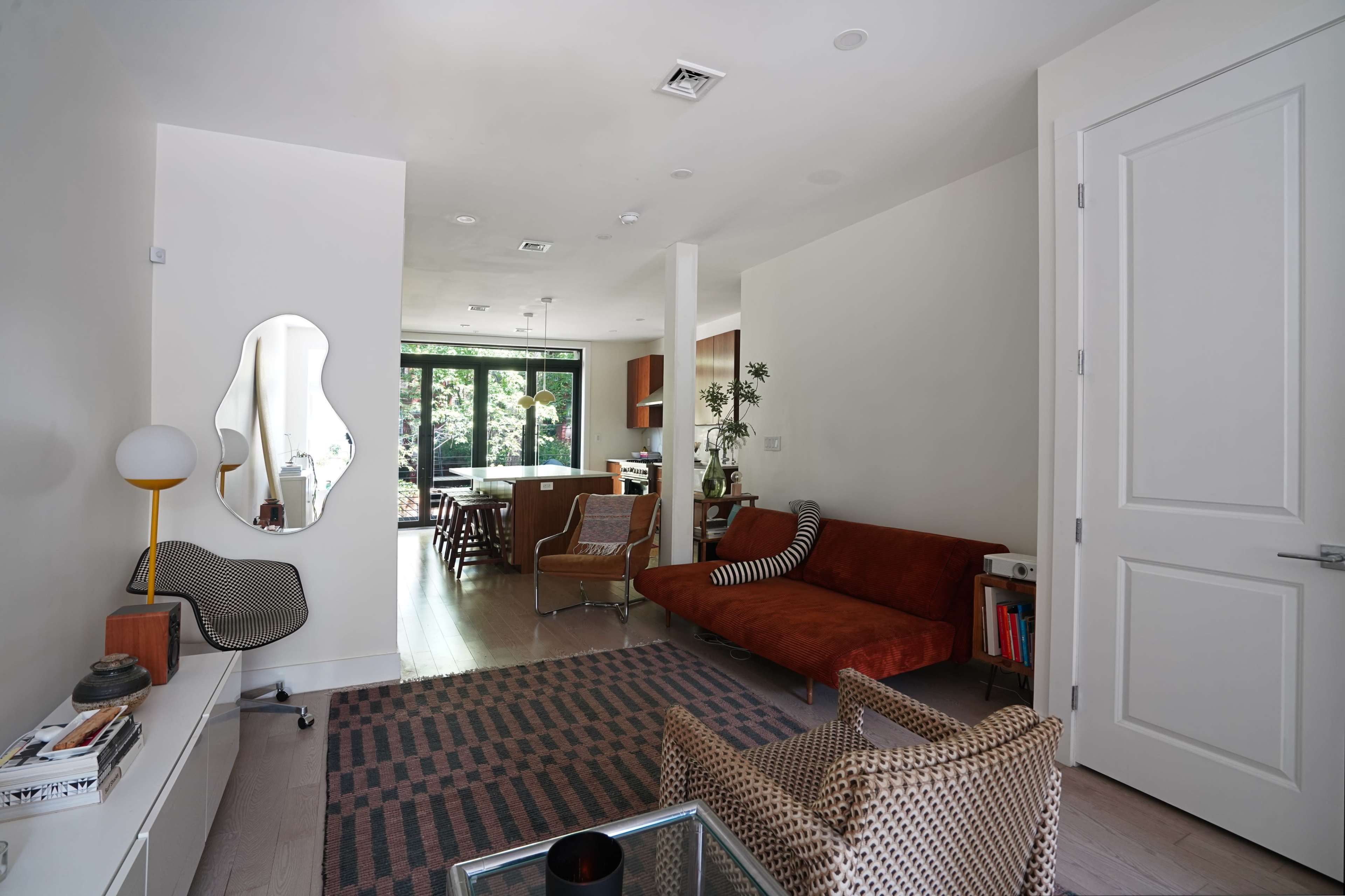 The image shows a modern living room with a brown sofa, two patterned chairs, a glass coffee table, and a view into a kitchen area through large windows.