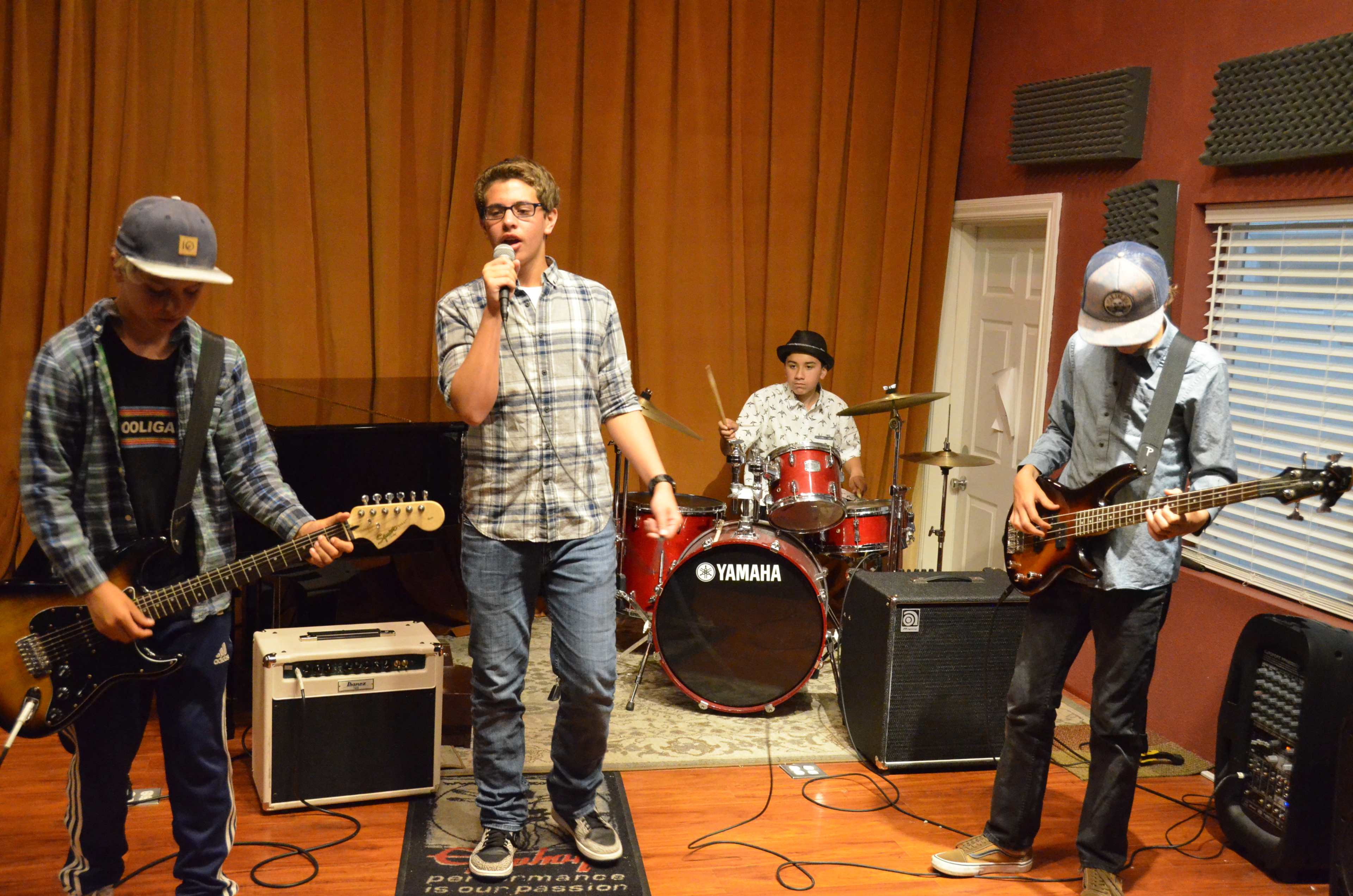 A group of four musicians performs in a studio, with one singer holding a microphone while the others play guitar, bass, and drums.