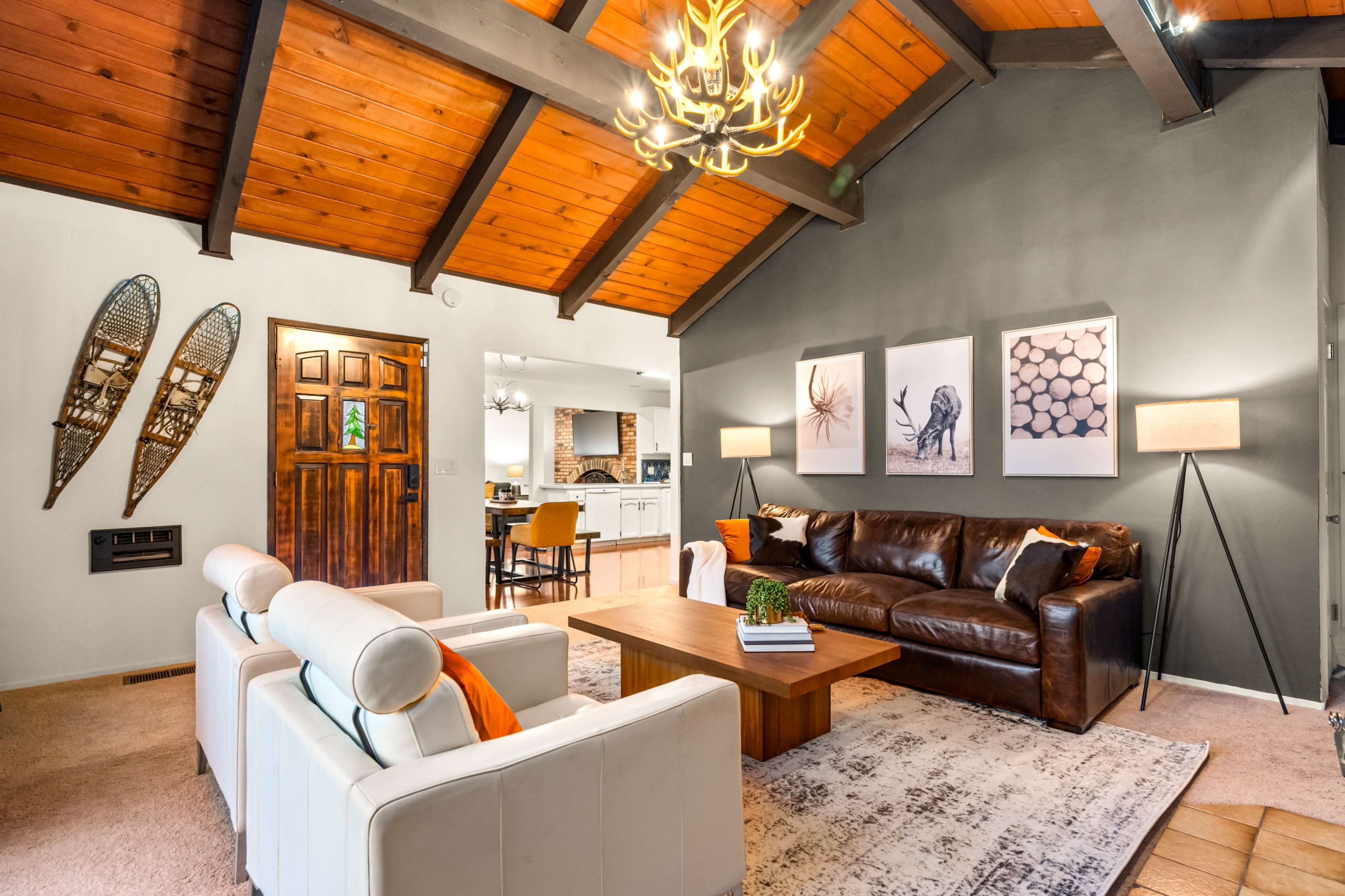 A spacious living room with a brown leather sofa, a wooden coffee table, and framed artwork on the walls, featuring exposed wooden beams and a chandelier overhead.